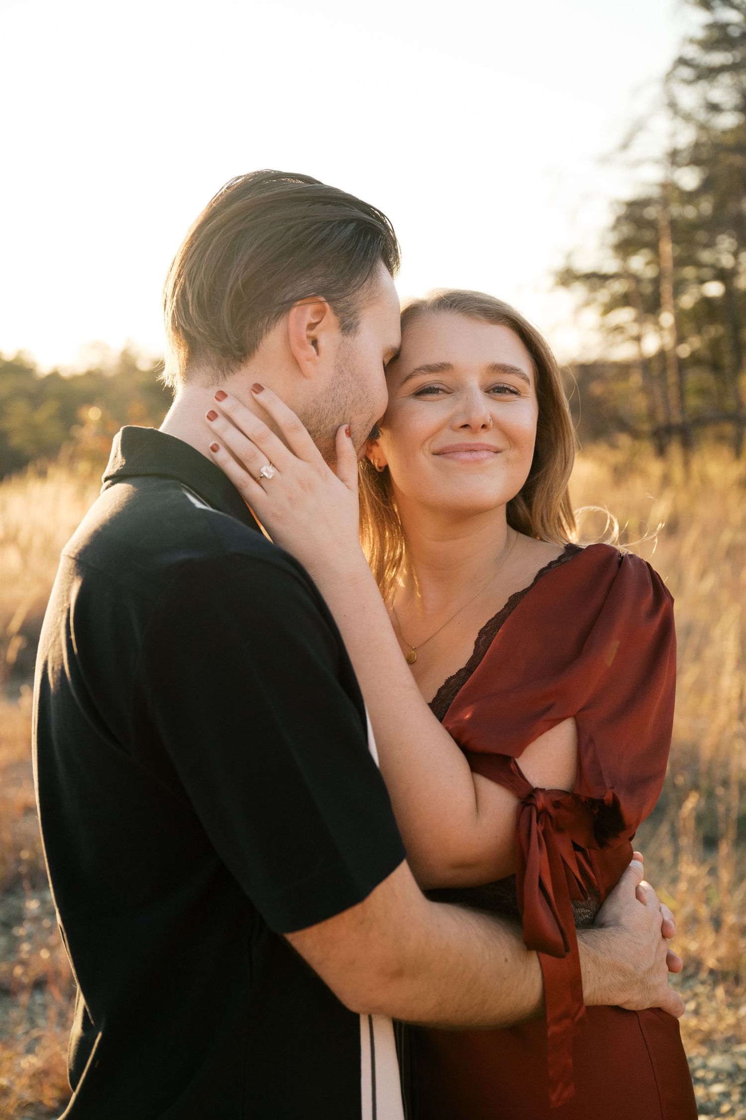 A couple embraces tenderly in a sunlit field at golden hour, with the woman in a burgundy dress smiling warmly.