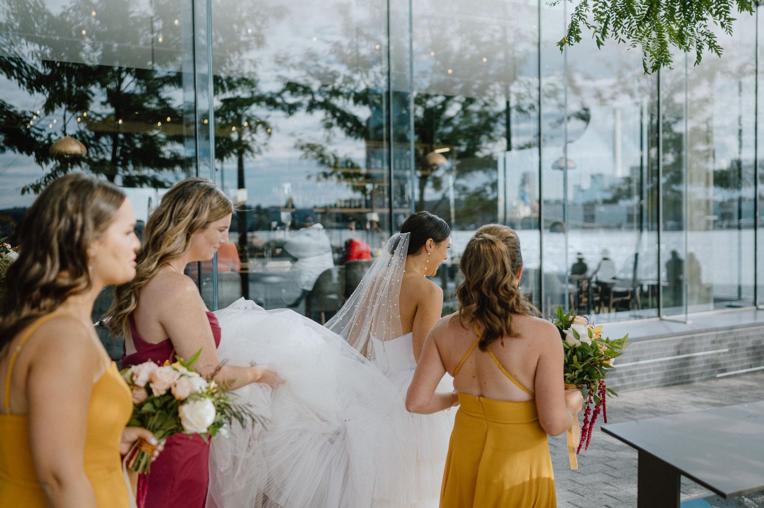 Bridesmaids in yellow dresses hold bouquets while standing behind white-clad bride near reflective glass windows.