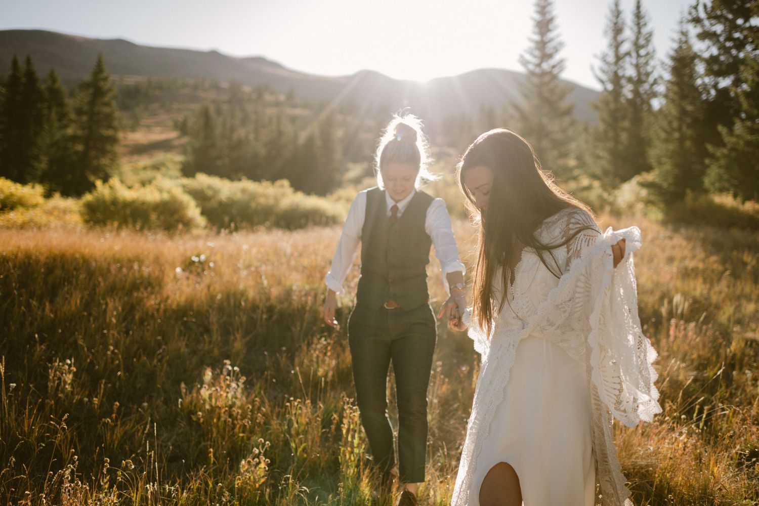Two people in wedding attire walking through a golden mountain meadow at sunset.