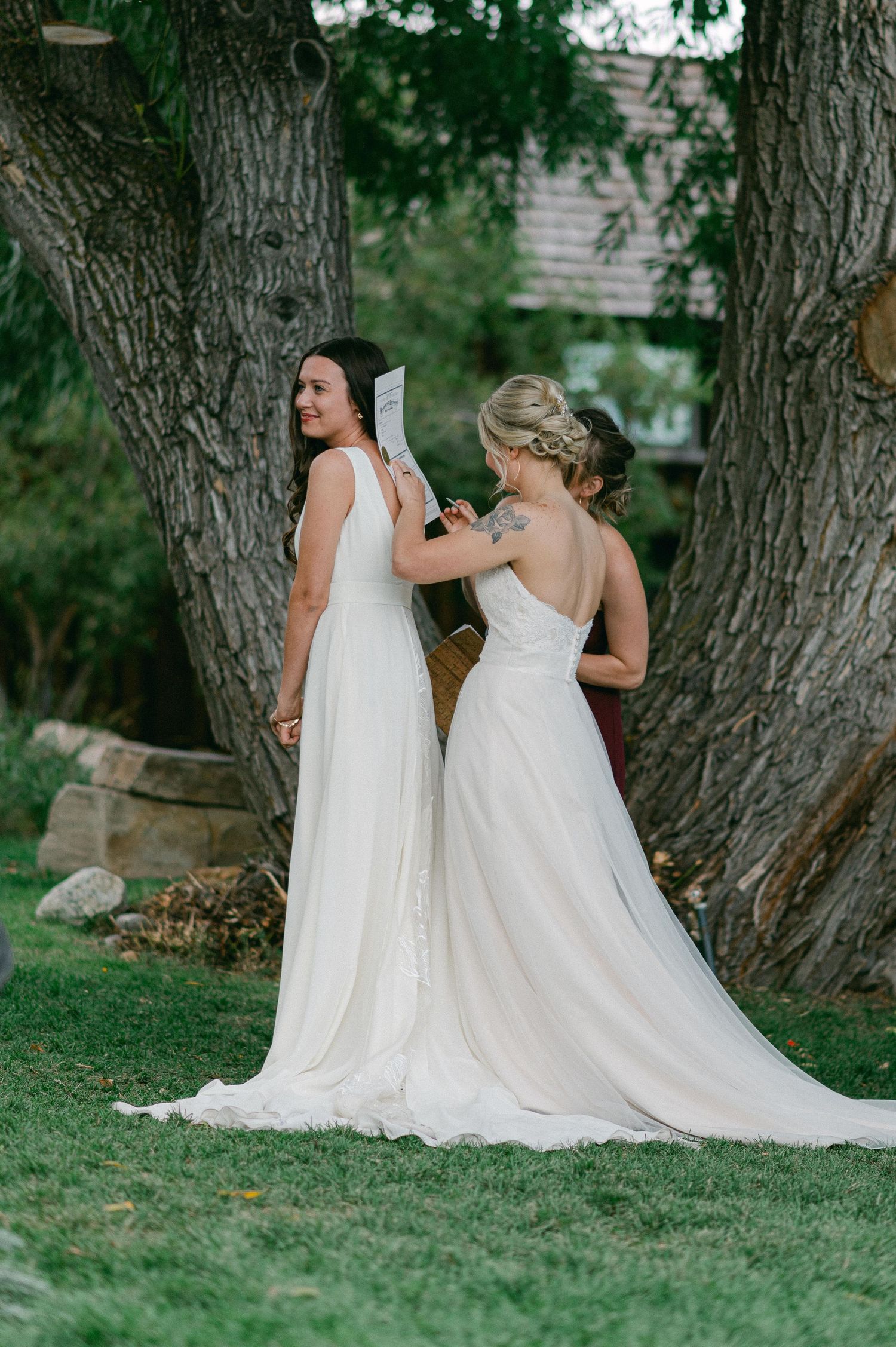 Two white wedding dresses photographed against the backdrop of mature trees in a garden setting.
