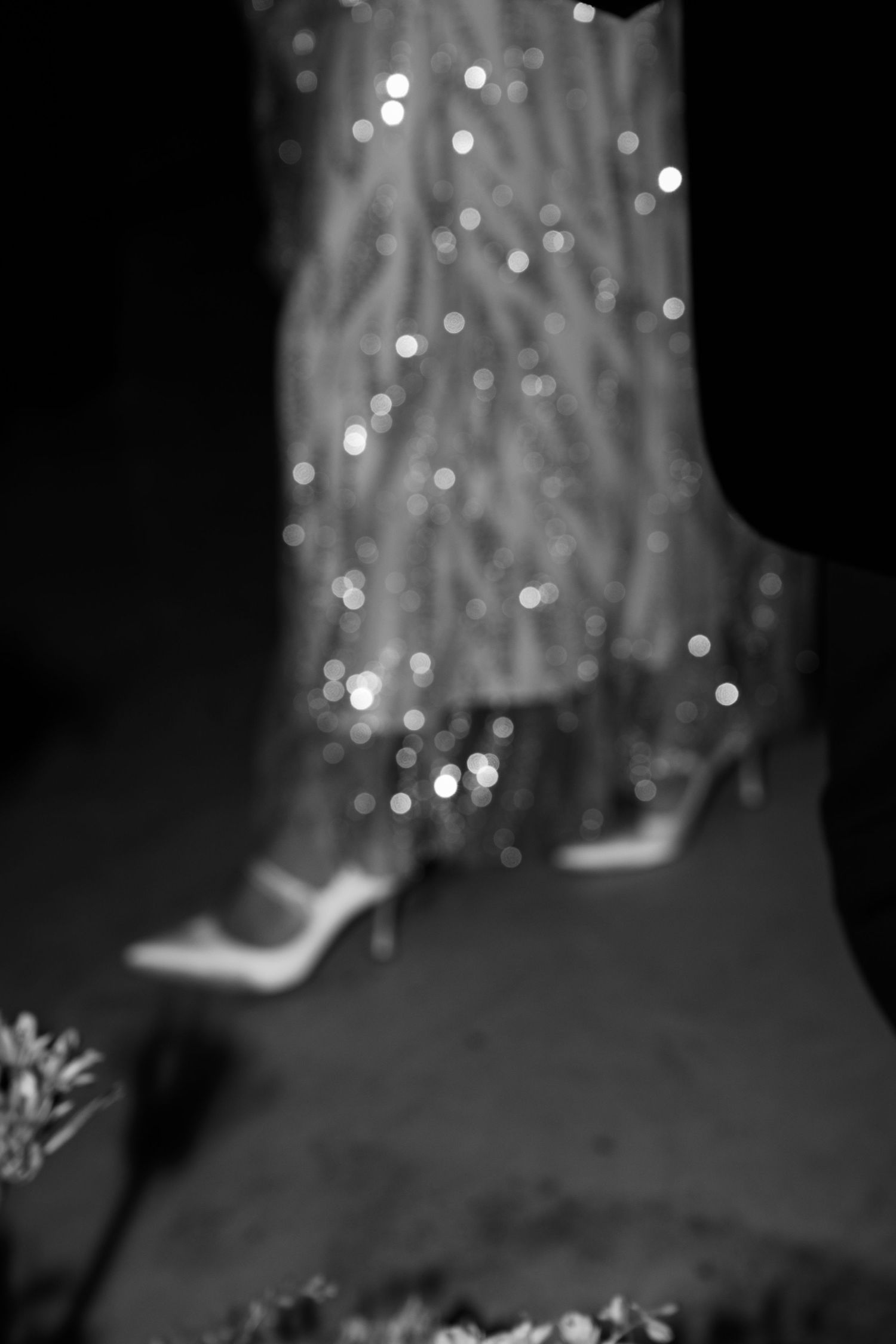 Close-up of sparkling bridal shoes and dress hem with bokeh lighting effects in black and white.