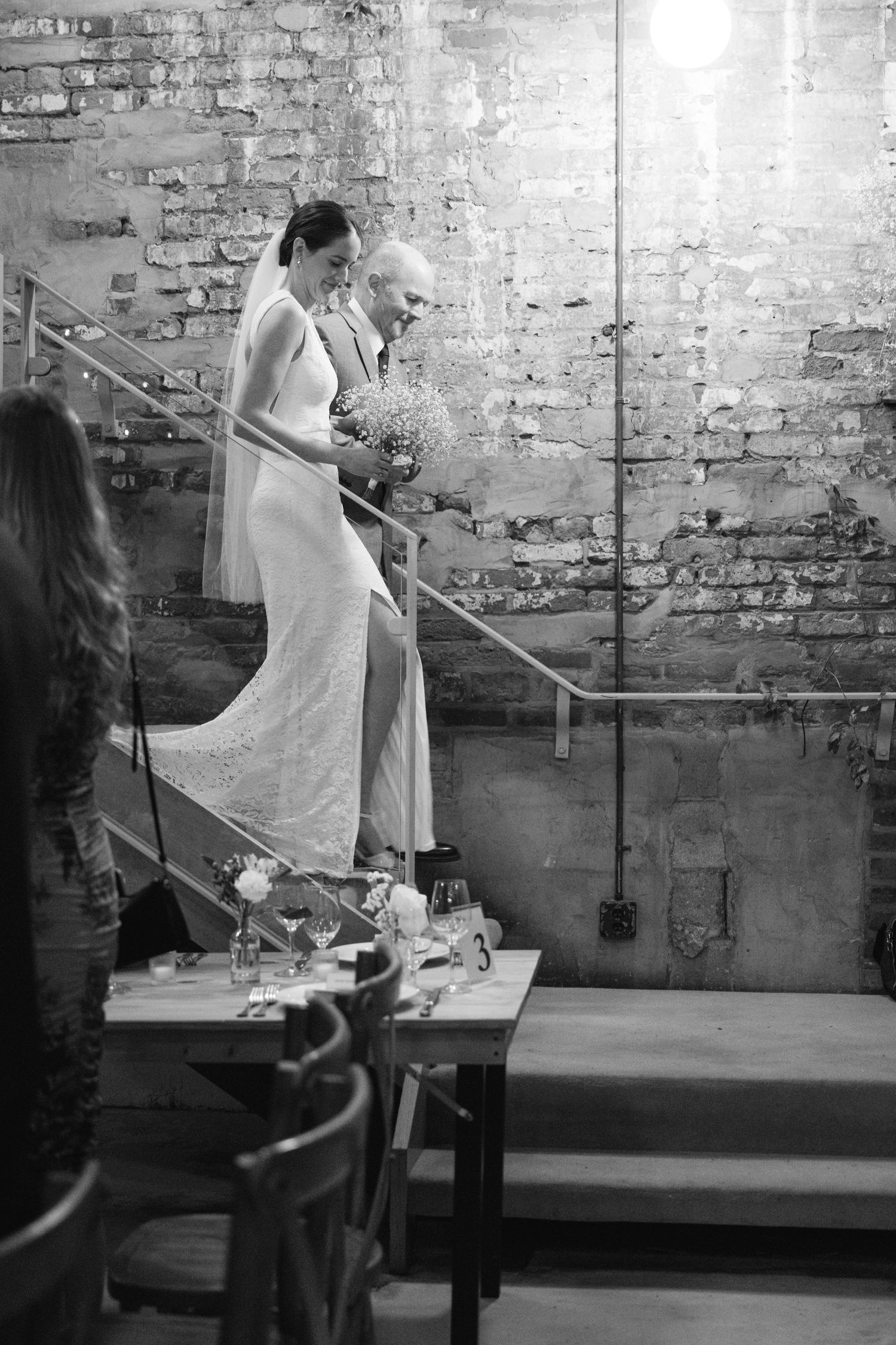 A bride descends stairs in an elegant white gown at an industrial-style venue with exposed brick walls and rustic decor.