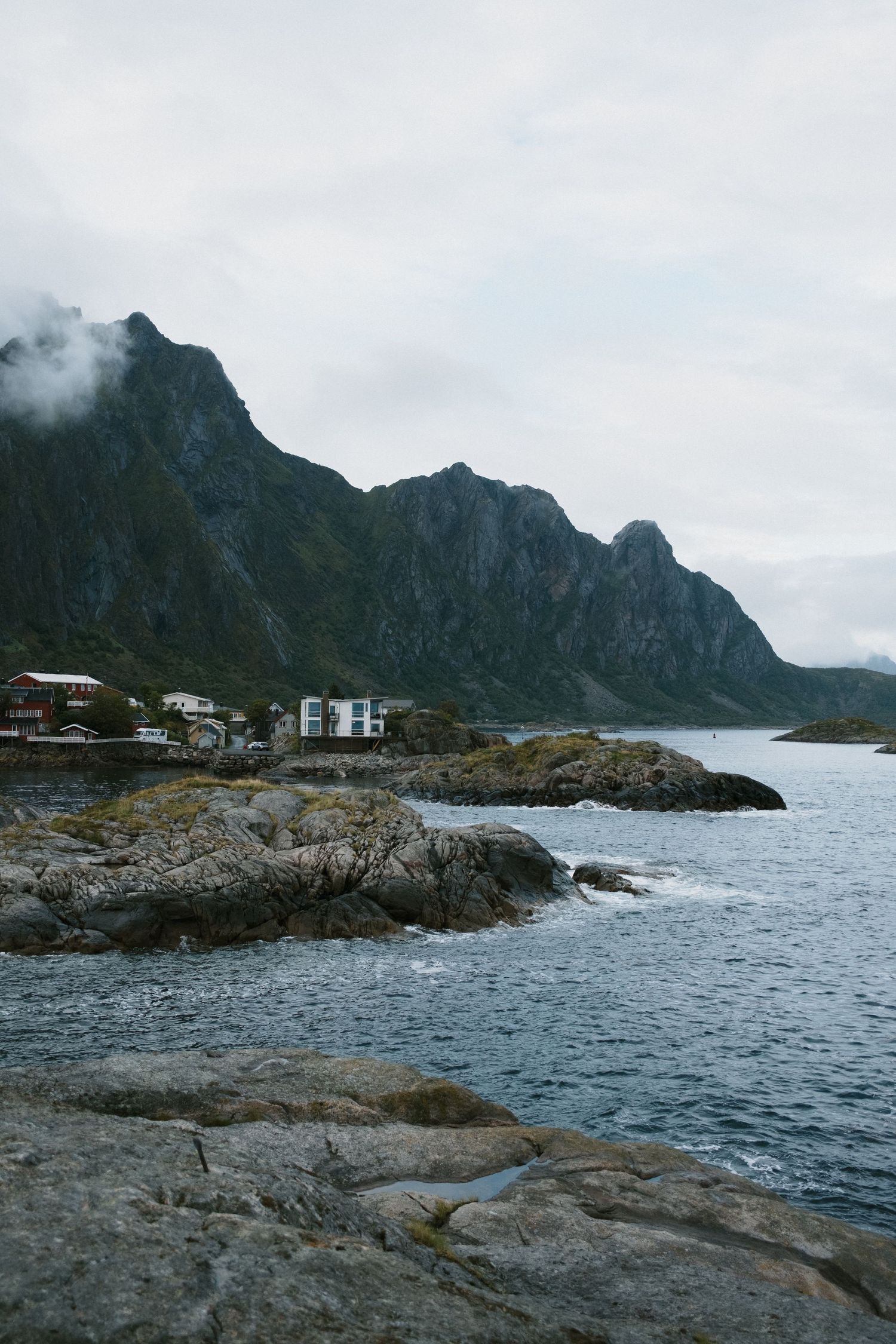 Rugged coastal landscape with houses perched on rocks beneath steep mountain cliffs.