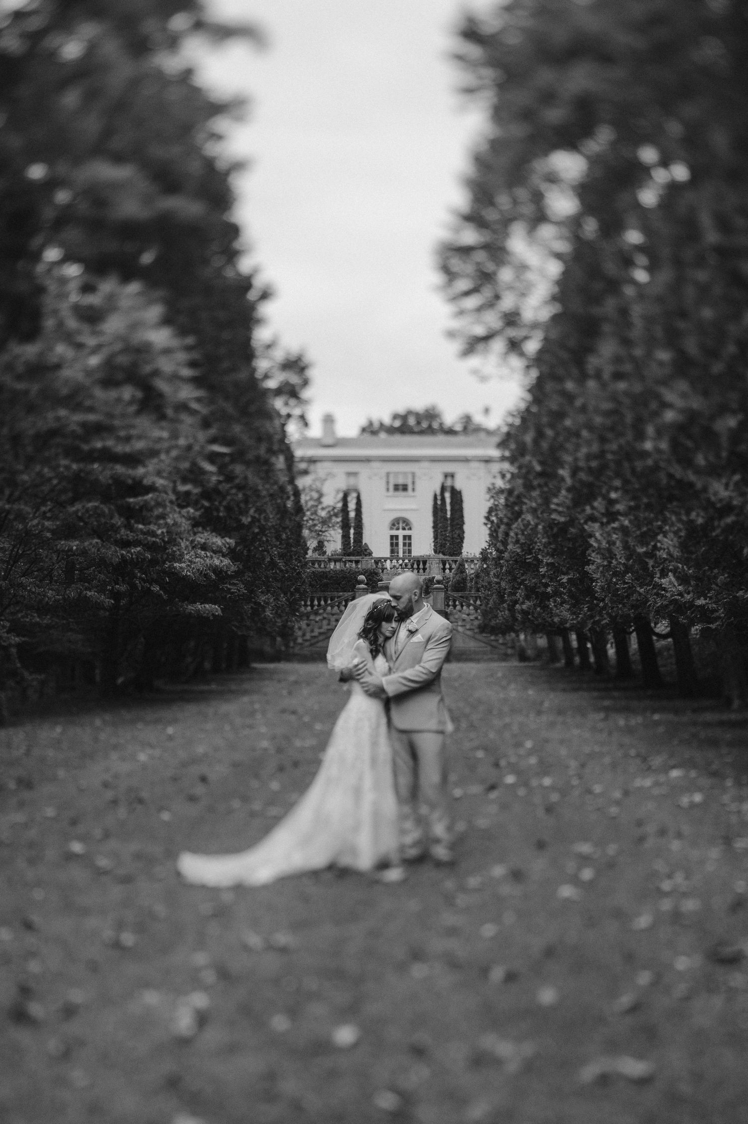 Romantic wedding couple embraces on a tree-lined path leading to an elegant white mansion in black and white.