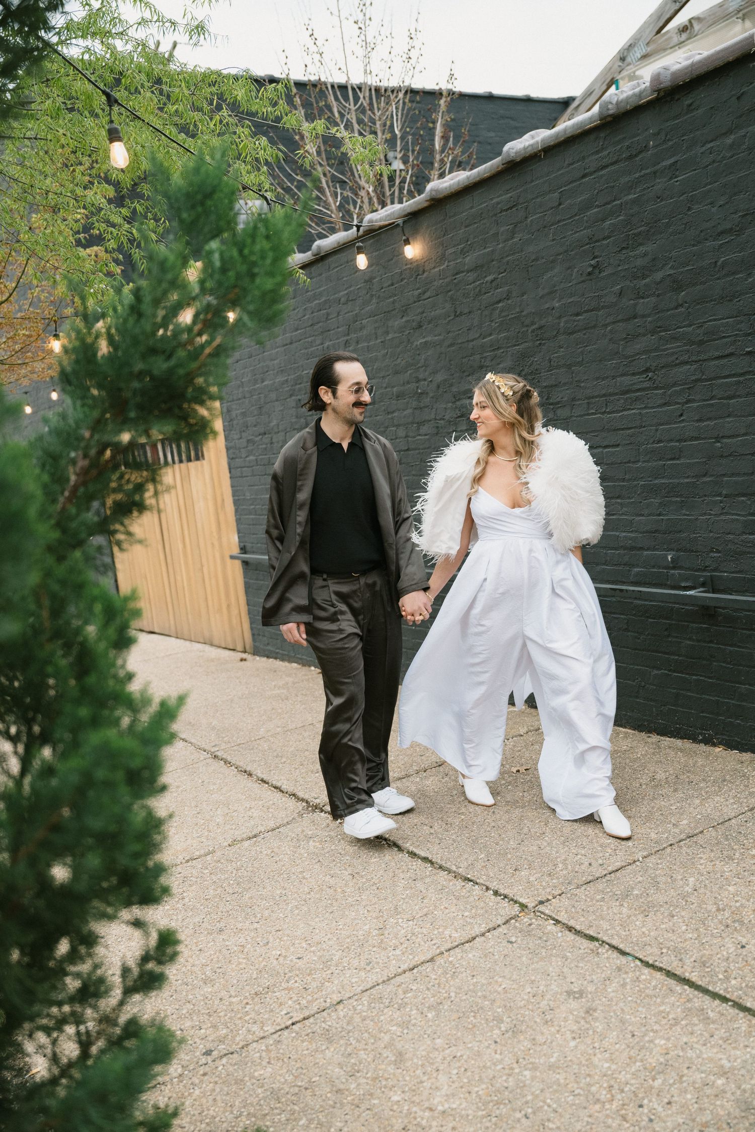 A couple wearing black and white outfits walk together on a sidewalk with string lights and greenery in an urban setting at Fadensonnen