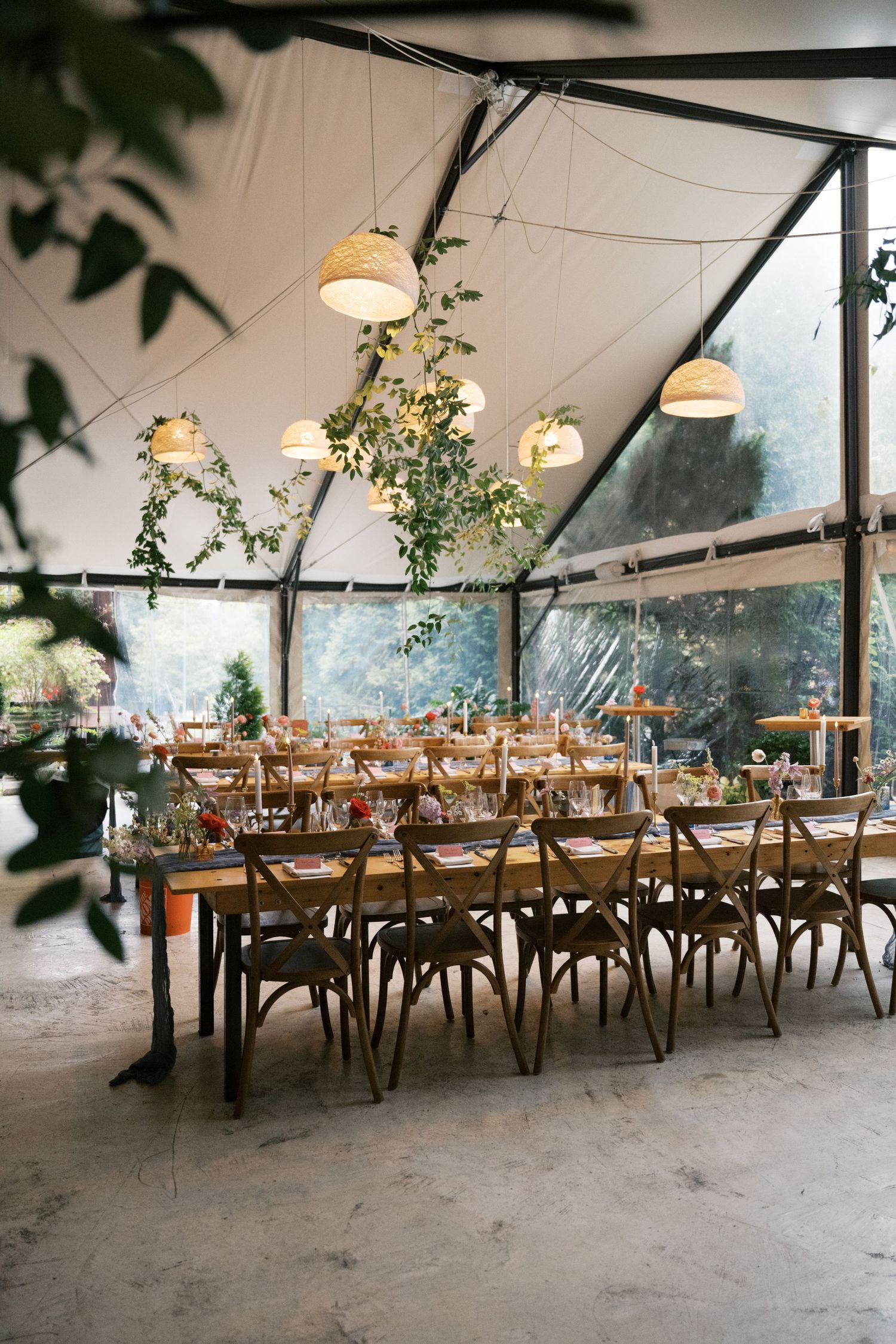 Elegant greenhouse wedding reception setup with rustic wooden tables, cross-back chairs, and hanging pendant lights.