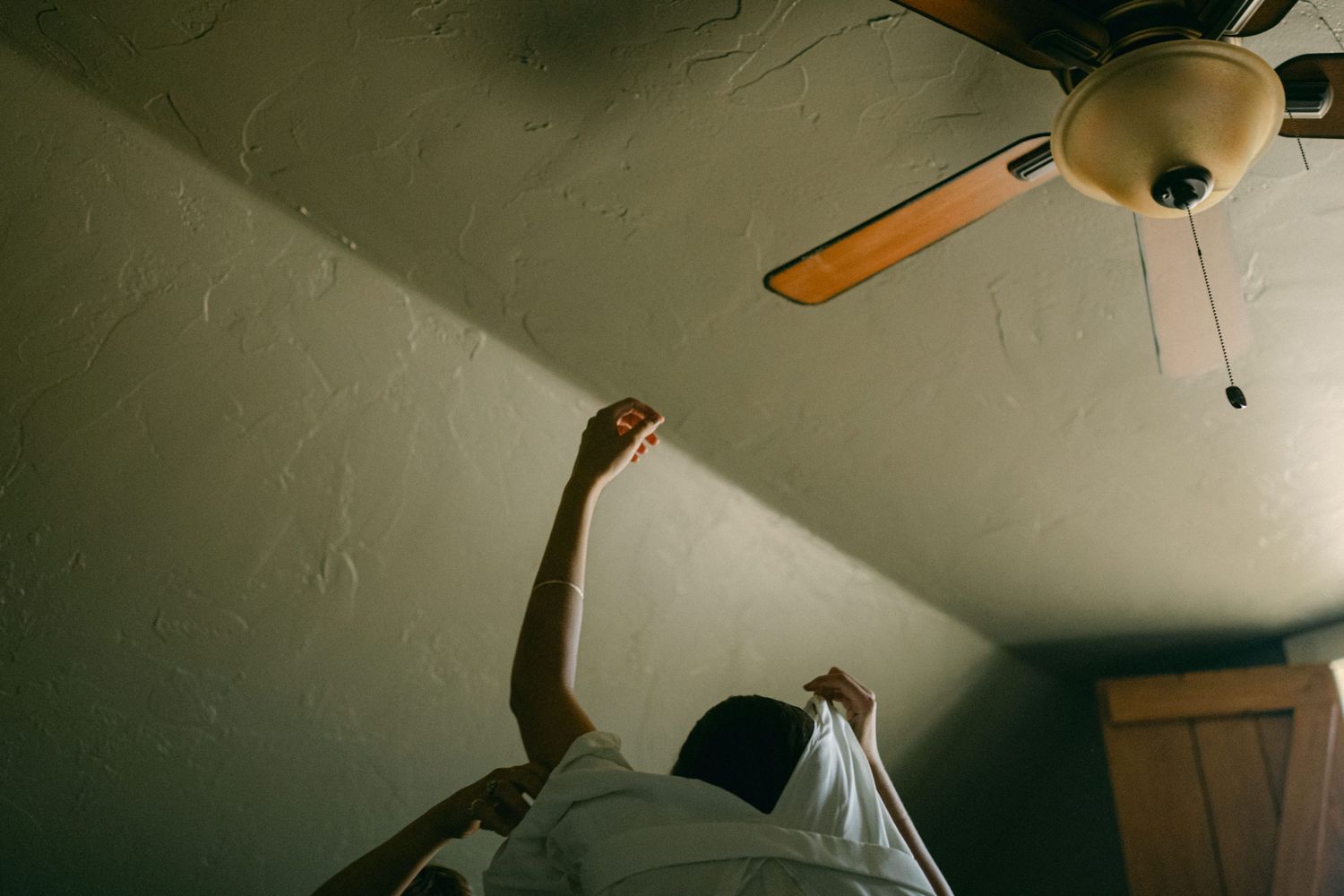White wedding dress hanging from ceiling fan in sunlit room.