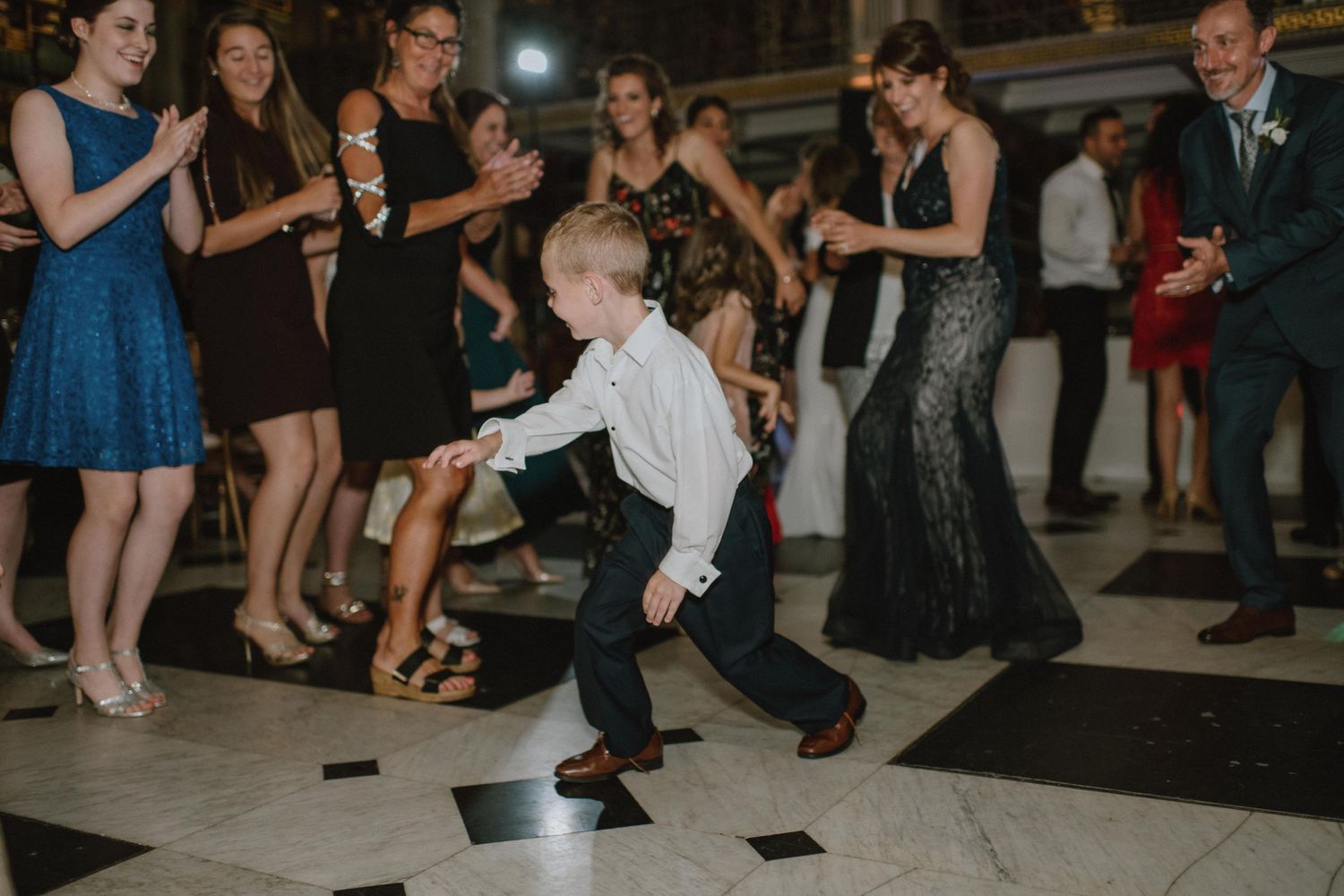 Young child shows off dance moves on checkerboard floor while wedding guests watch and cheer.
