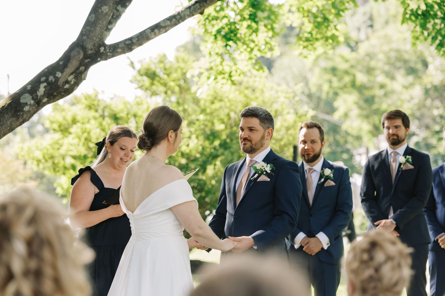 A wedding ceremony takes place outdoors under a tree canopy with attendees in formal attire.