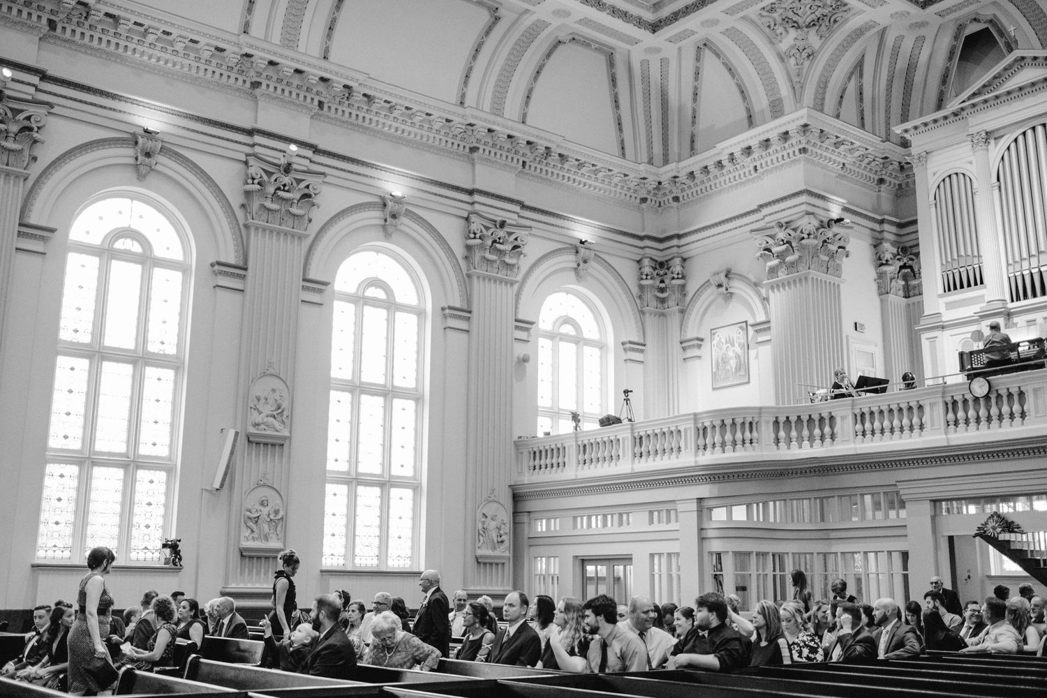 Black and white photo of wedding guests seated in an ornate church with tall arched windows.
