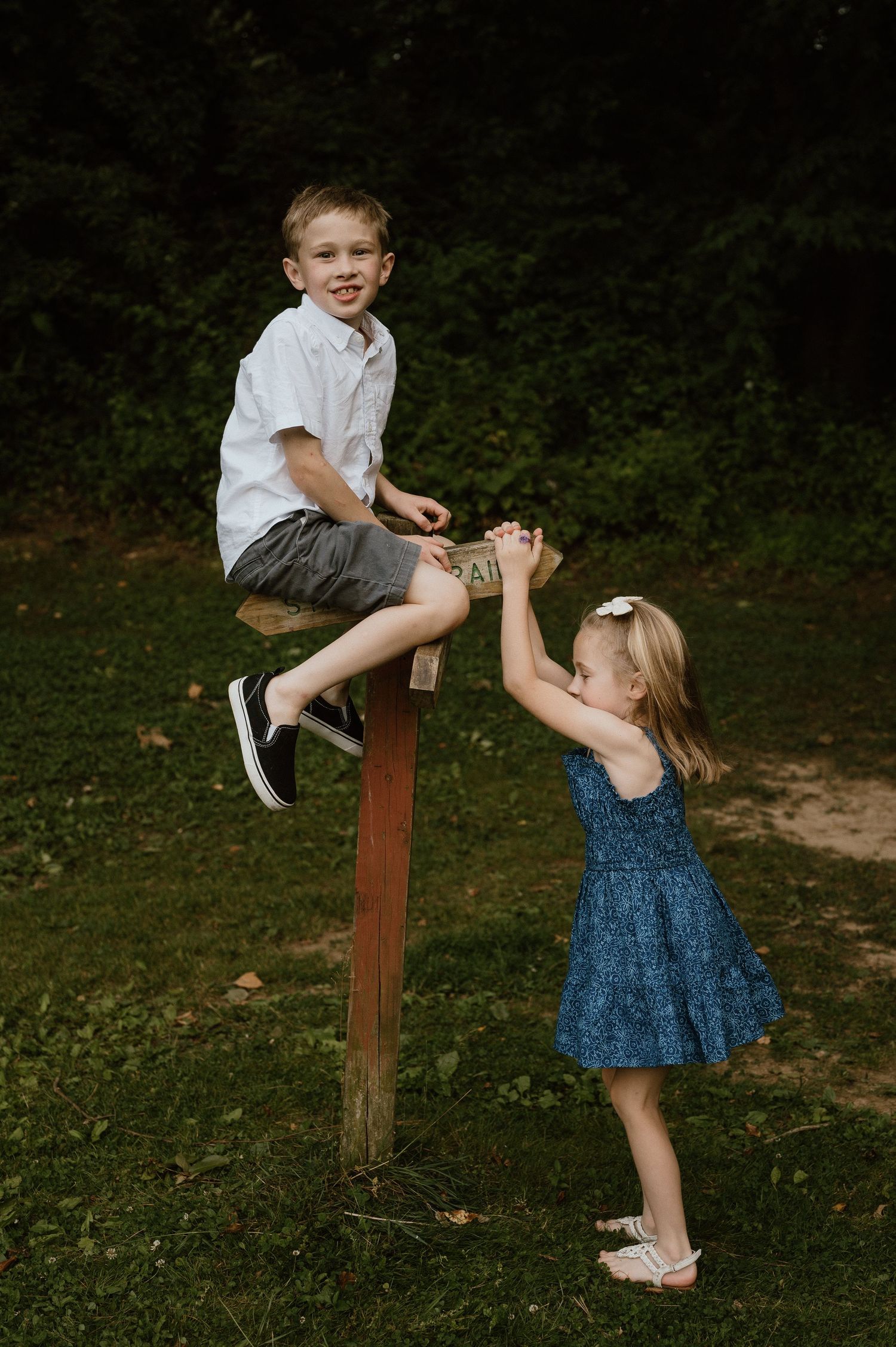 Two children playing outdoors, one sitting on wooden post while other reaches up.