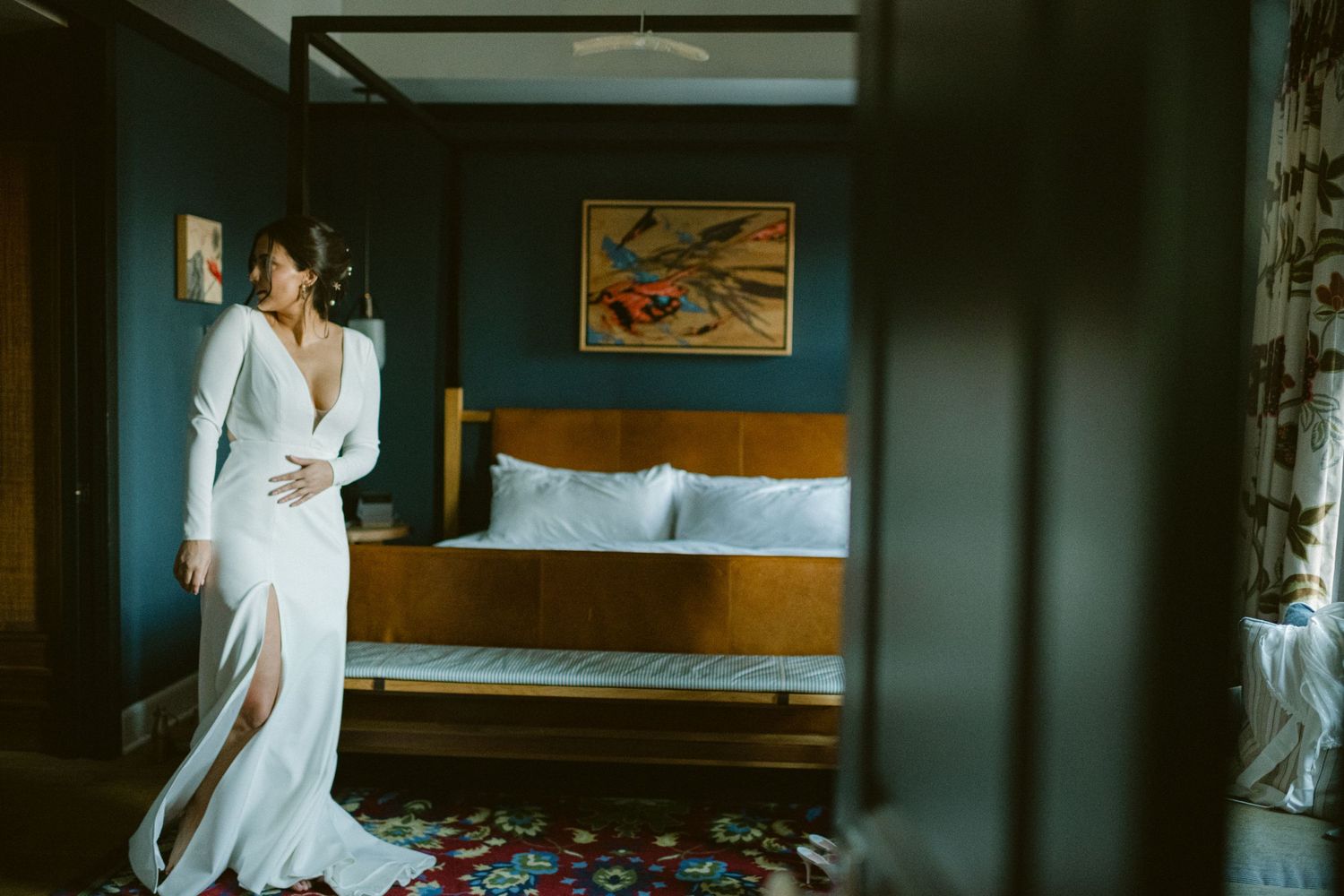 Elegant white wedding dress being fitted in a modern hotel room with blue walls and wooden furniture.