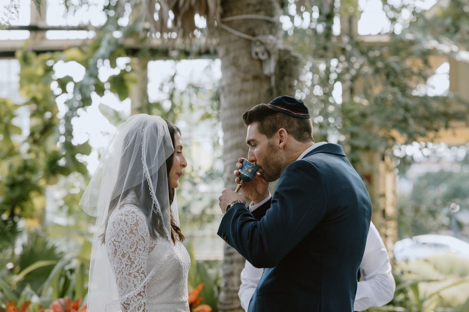 Groom kisses bride's hand during Jewish wedding ceremony in greenhouse.
