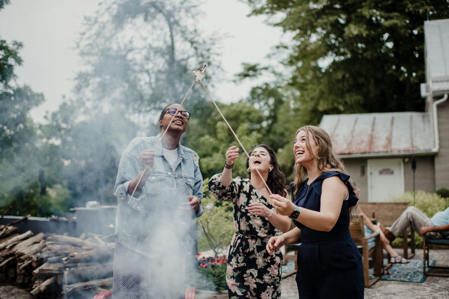 People laugh while gathered around a smoking fire pit in a backyard.