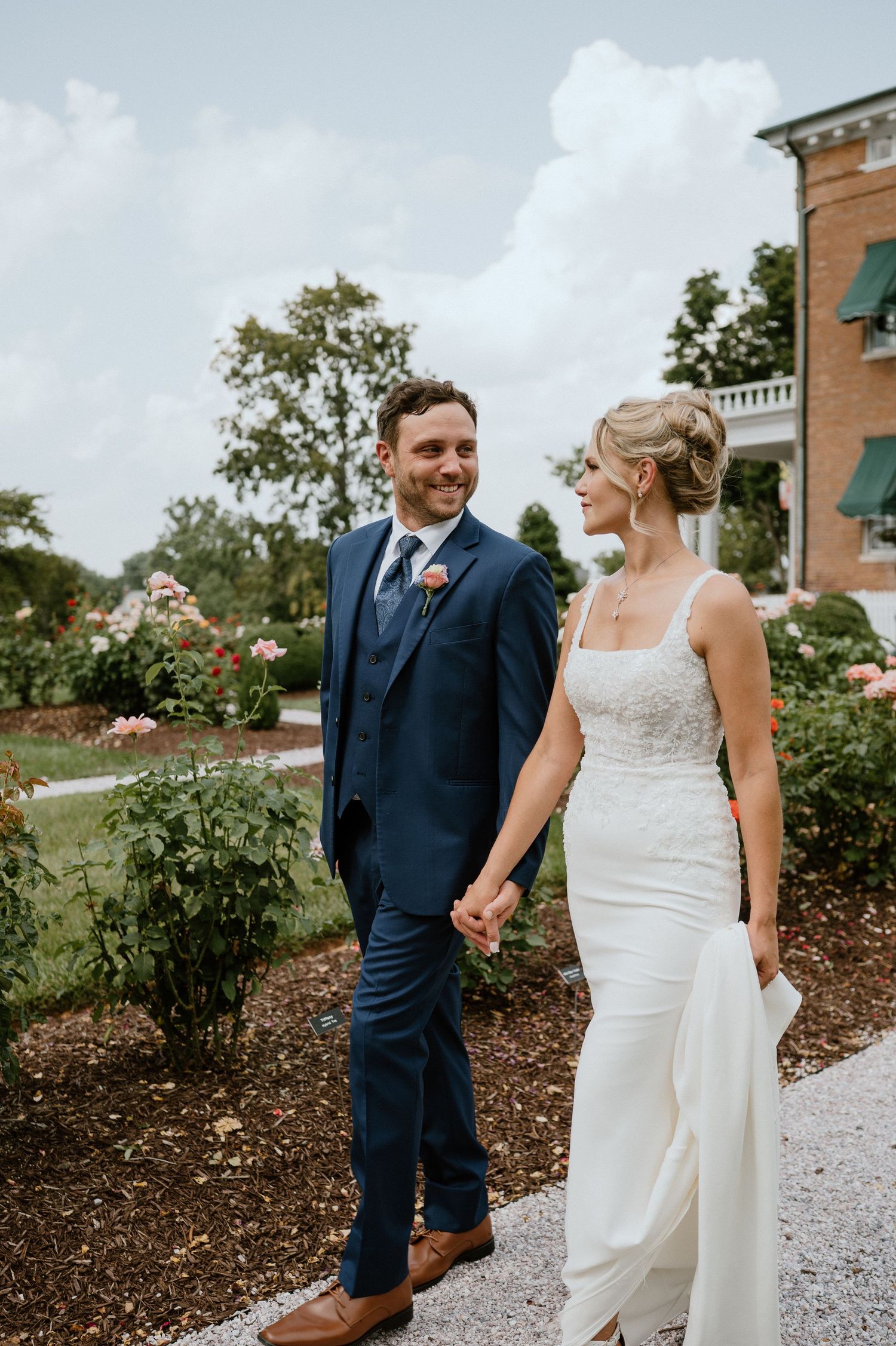 Newlywed couple walks hand in hand through a garden path wearing a navy suit and white wedding gown.