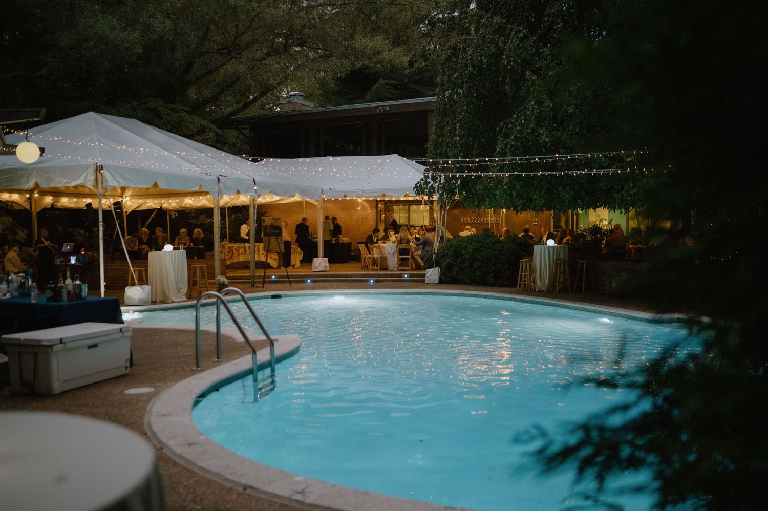 An illuminated pool area with string lights and white tents set up for an evening outdoor event.