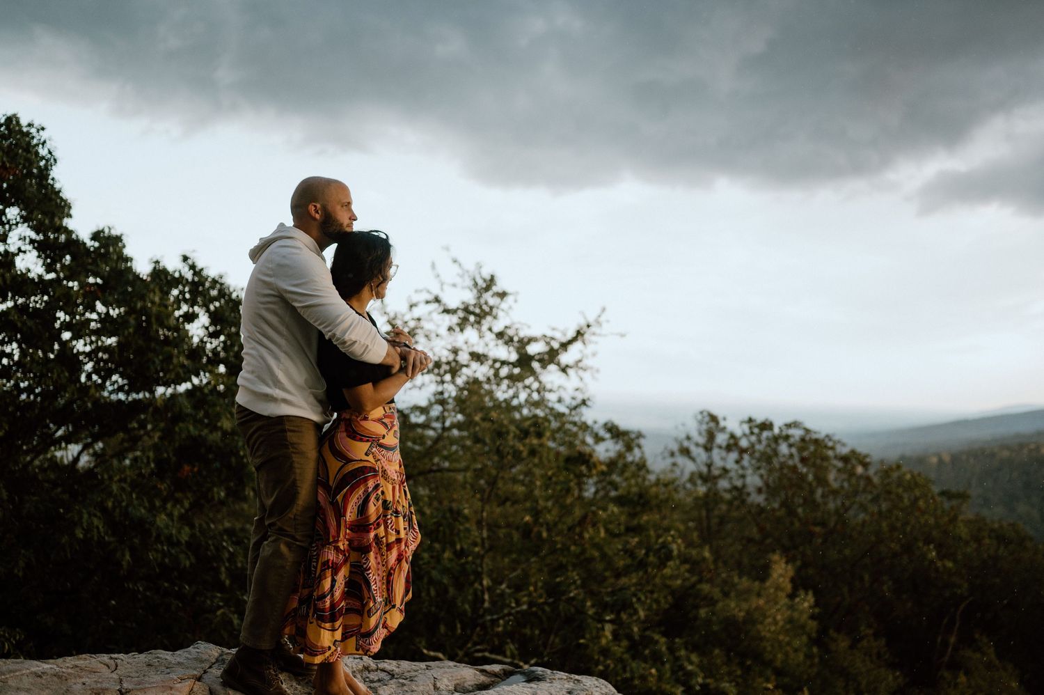 A couple embraces on a mountaintop against stormy skies and rolling hills in the distance.