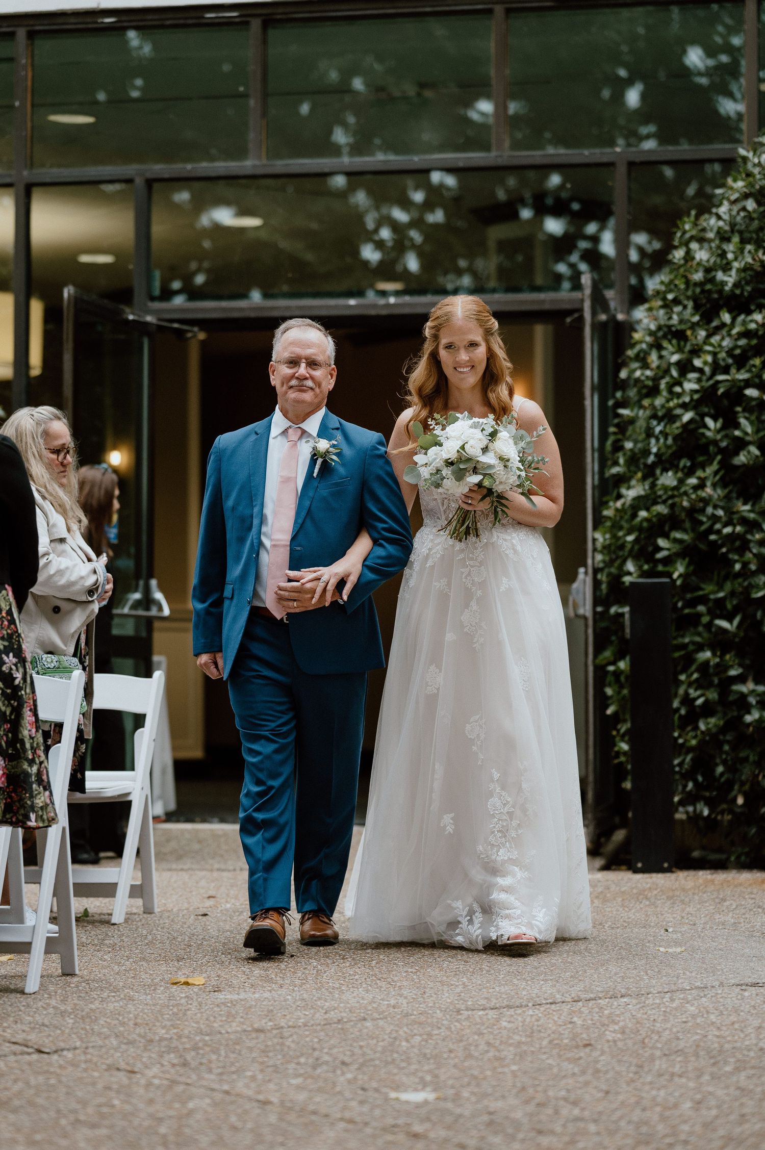 Bride in lace gown walks down aisle with escort at modern venue entrance.