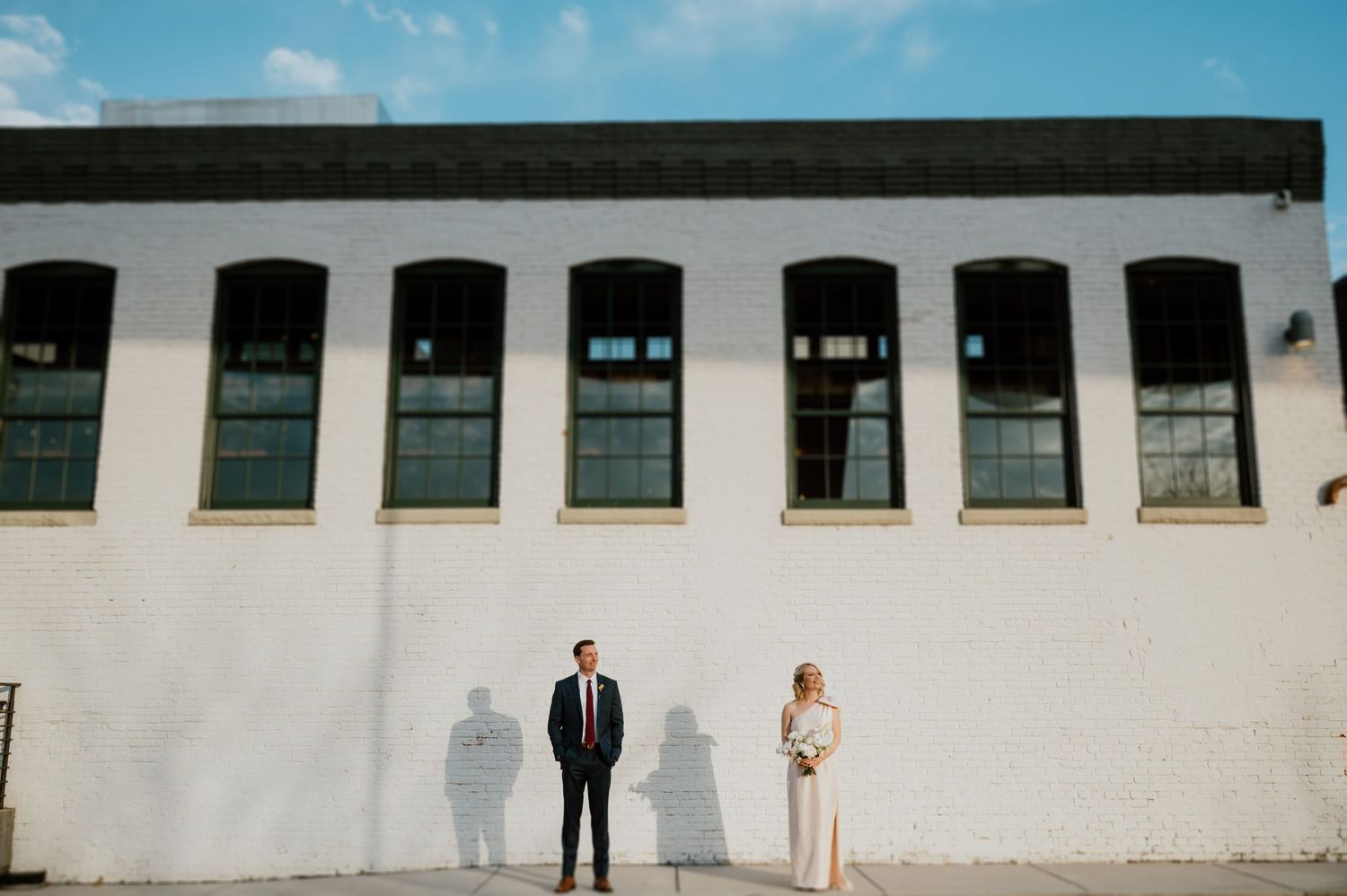 Bride and groom pose against a white industrial building with windows.