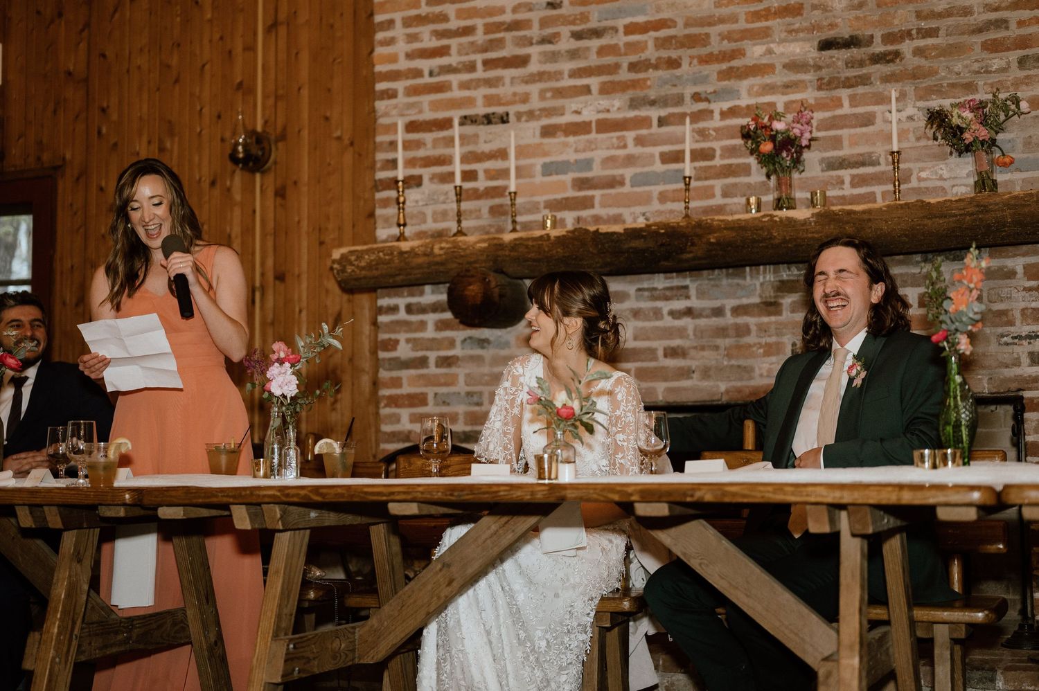 Wedding guests sharing laughter at a rustic reception with exposed brick walls and wooden tables.
