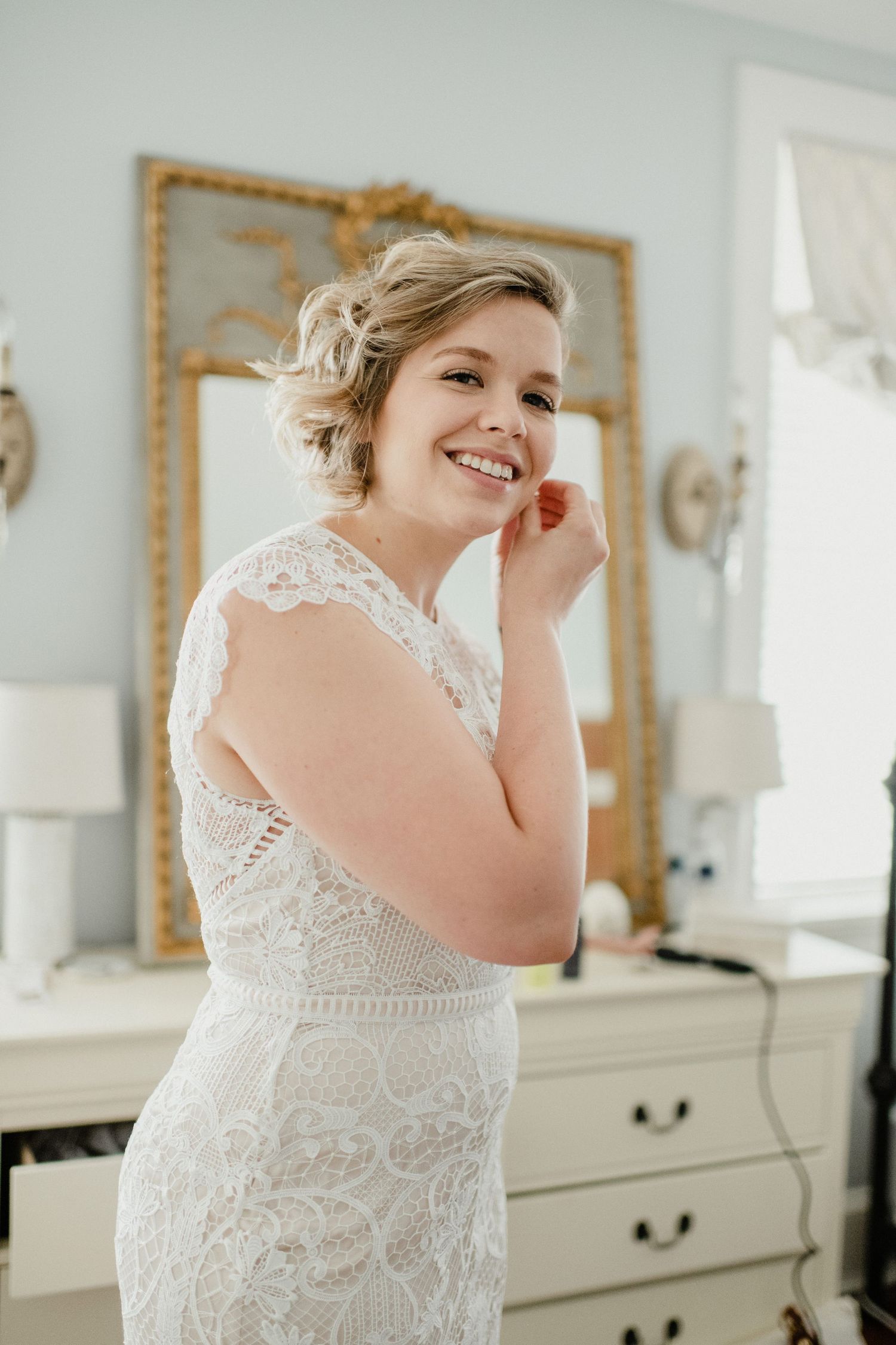 A bride wearing a detailed lace wedding dress smiles while getting ready in a bright room with a gold-framed mirror.