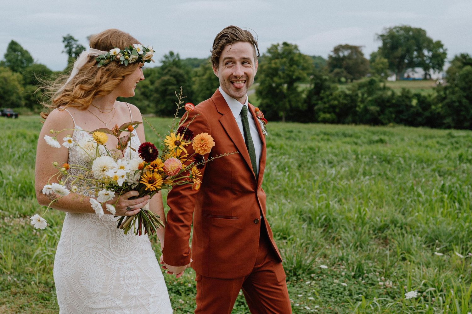 Wedding couple poses in green field with sunflower bouquet and rusty orange suit.