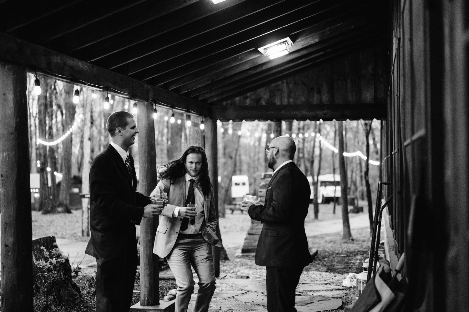 Black and white photo of three people chatting under a covered porch in the woods.