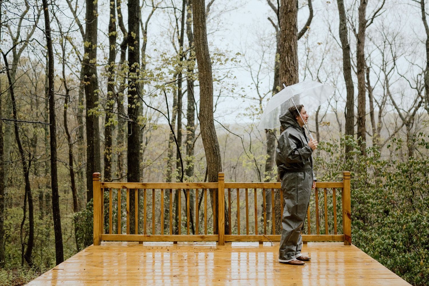 Figure in winter clothing stands on wet wooden deck surrounded by bare trees.