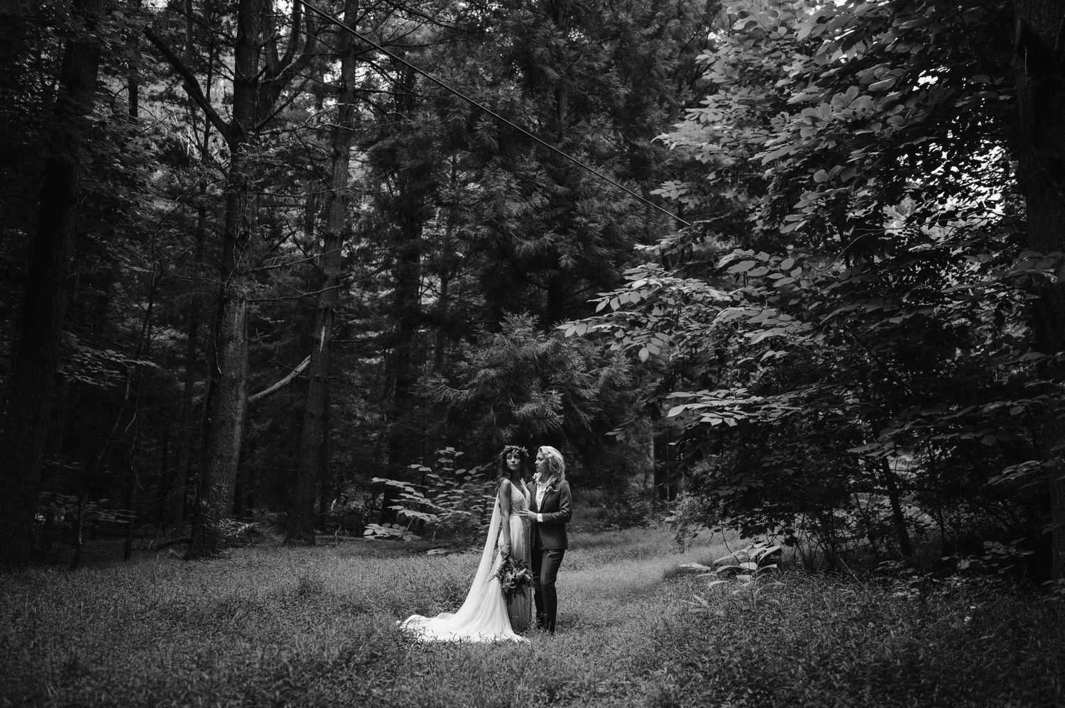 A black and white wedding portrait of a couple standing in a serene woodland clearing.