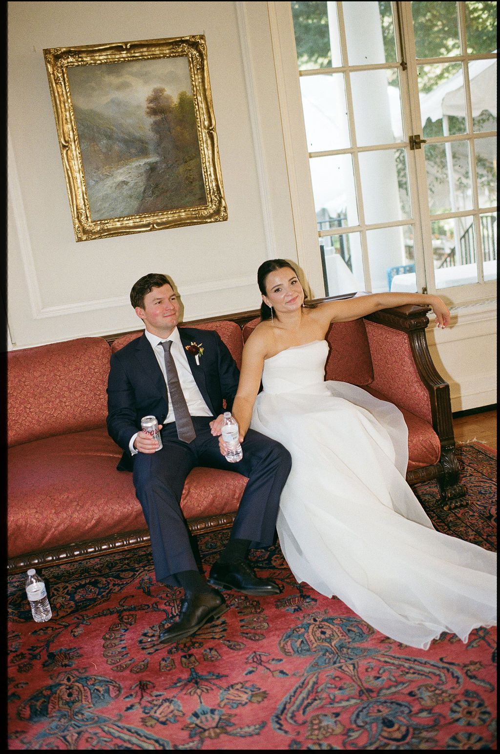 Wedding couple relaxing on an elegant red sofa in a formal room with ornate gold frames.