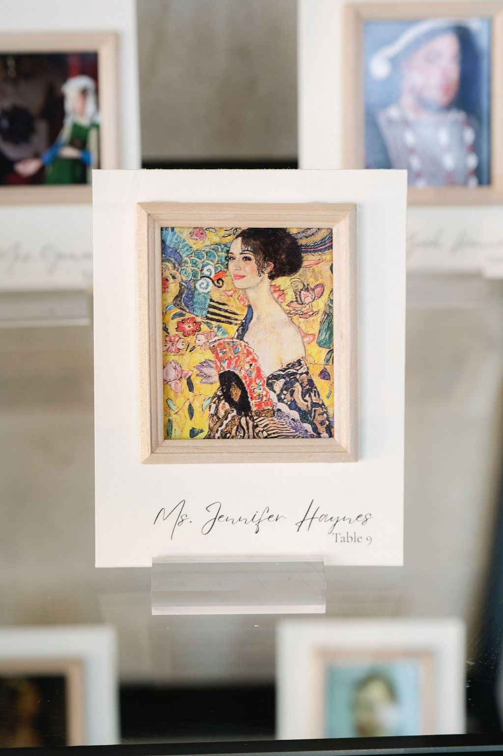 A gallery wall displays framed artwork including colorful Klimt-style paintings in white frames against a light background.