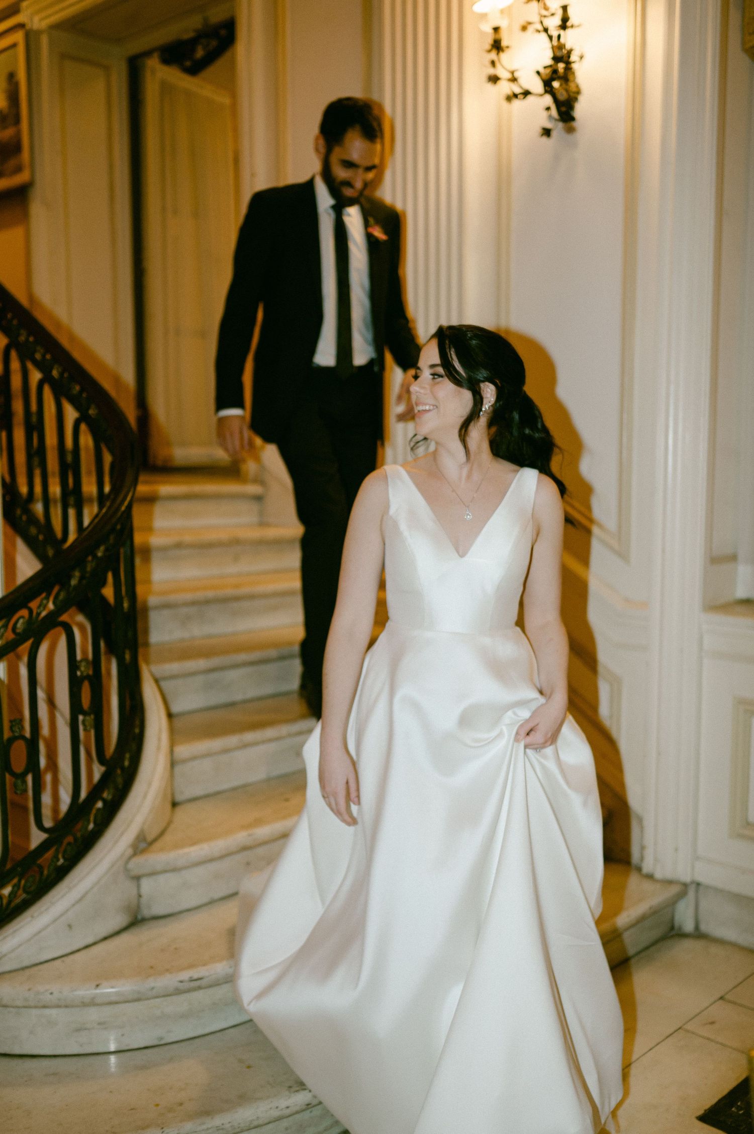 A bride in an elegant white gown walks through a grand hallway with classical architecture.