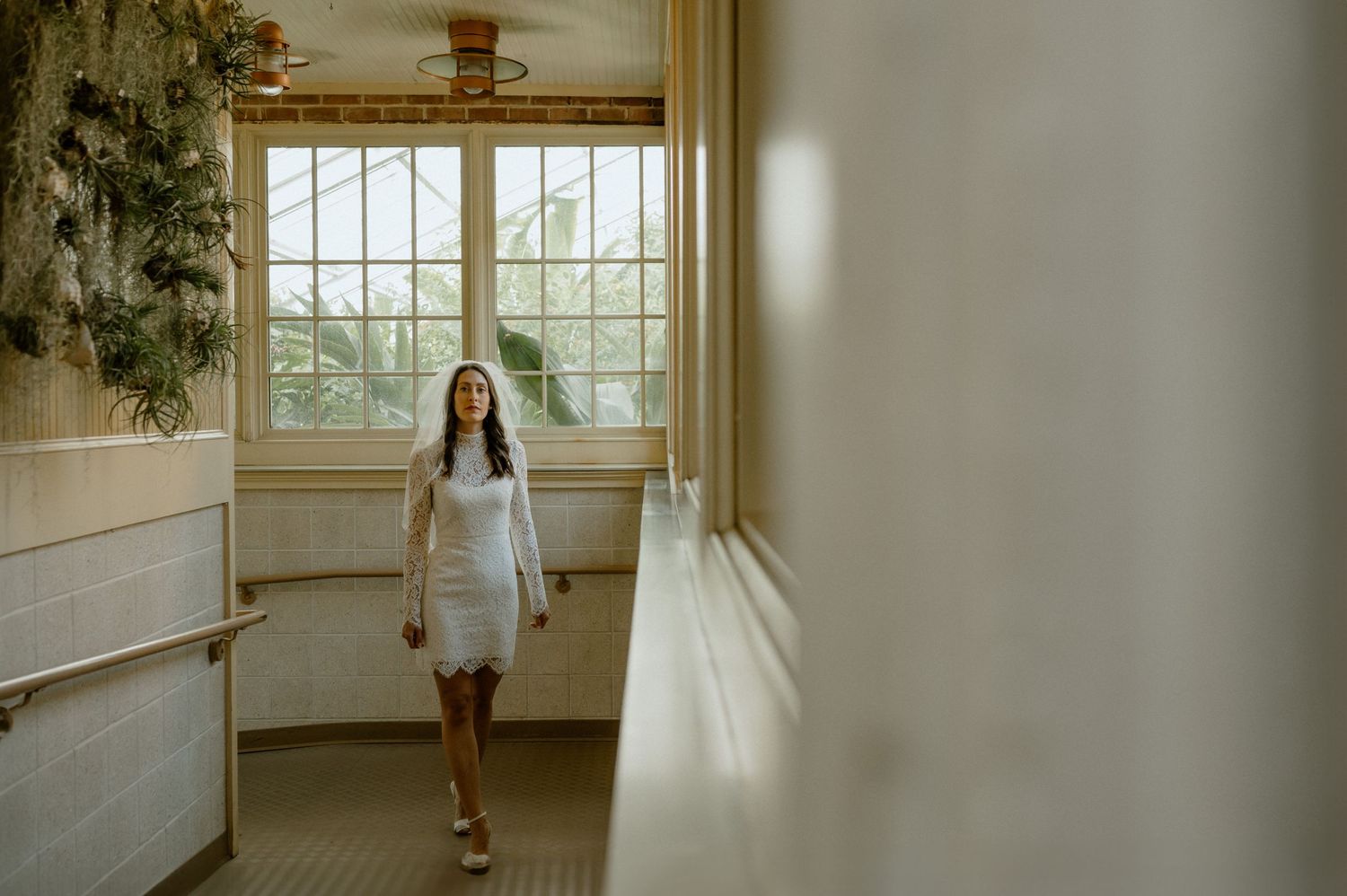 A figure in a white dress walks down a sunlit hallway with large windows and indoor plants.