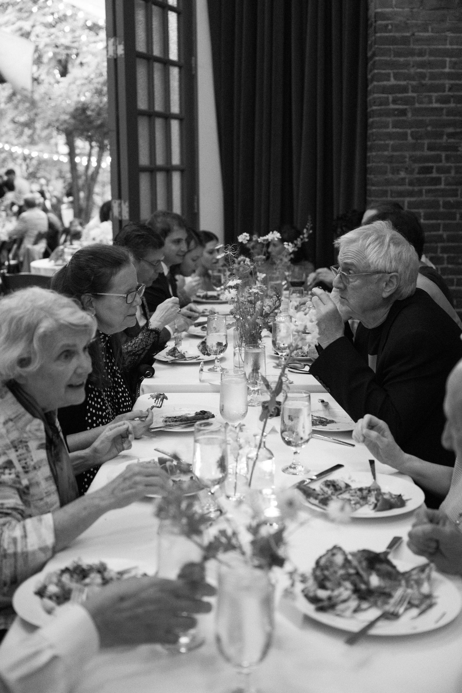A black and white photograph of people dining at a long table with flowers and plates, captured in an intimate setting.