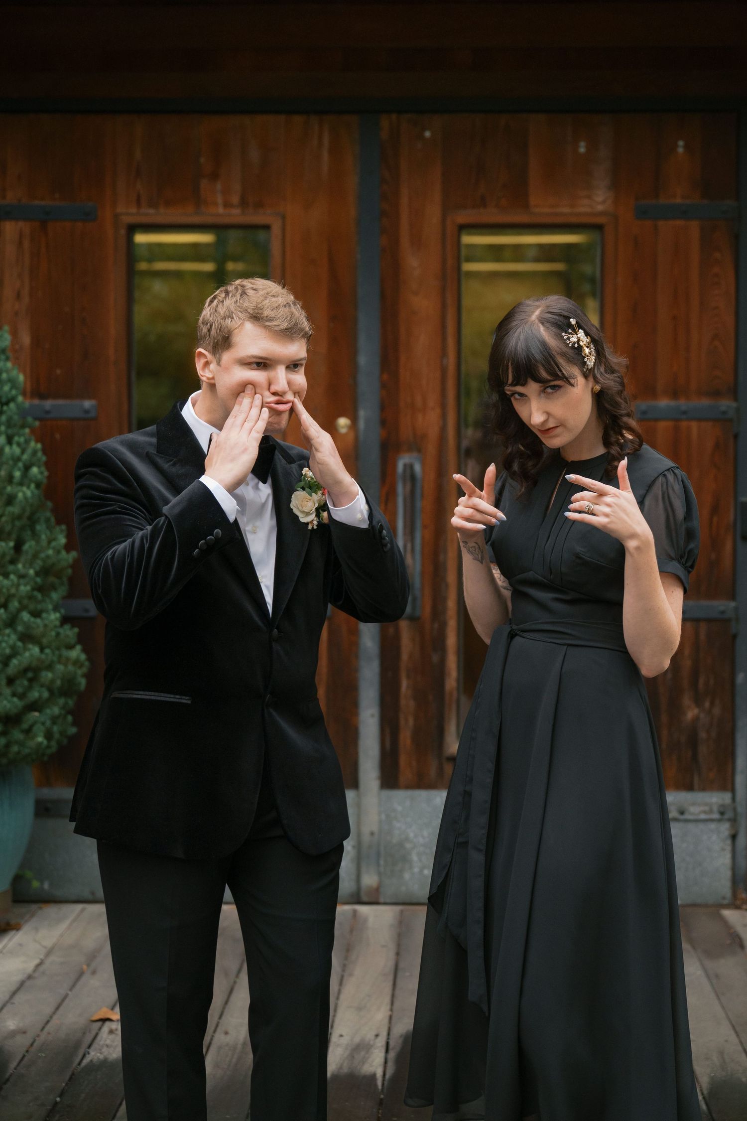 Two people in formal attire make playful gestures in front of wooden doors.