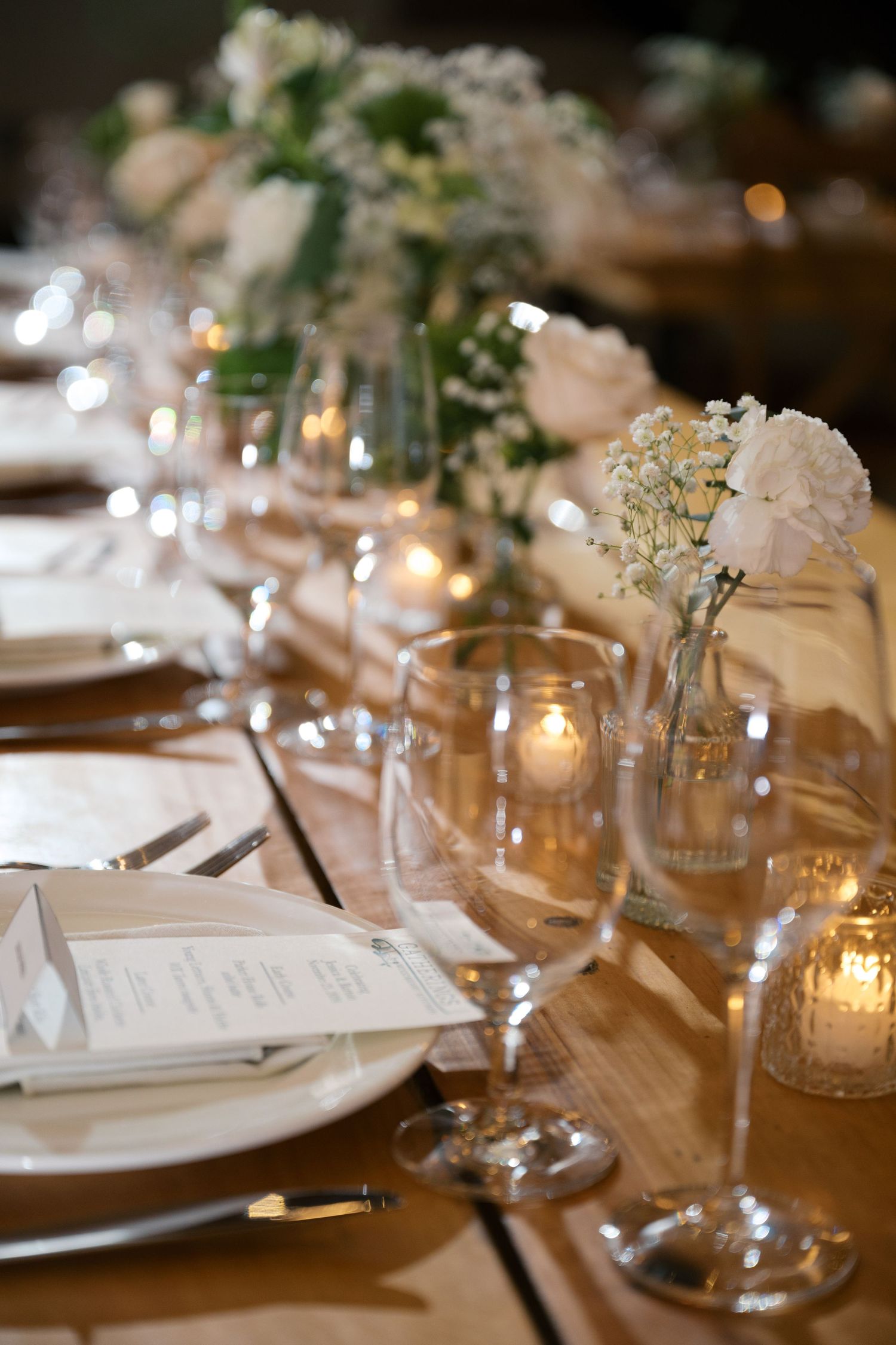 Elegant wedding table setting with candlelit mason jars, white flowers, and place settings on rustic wooden surface.