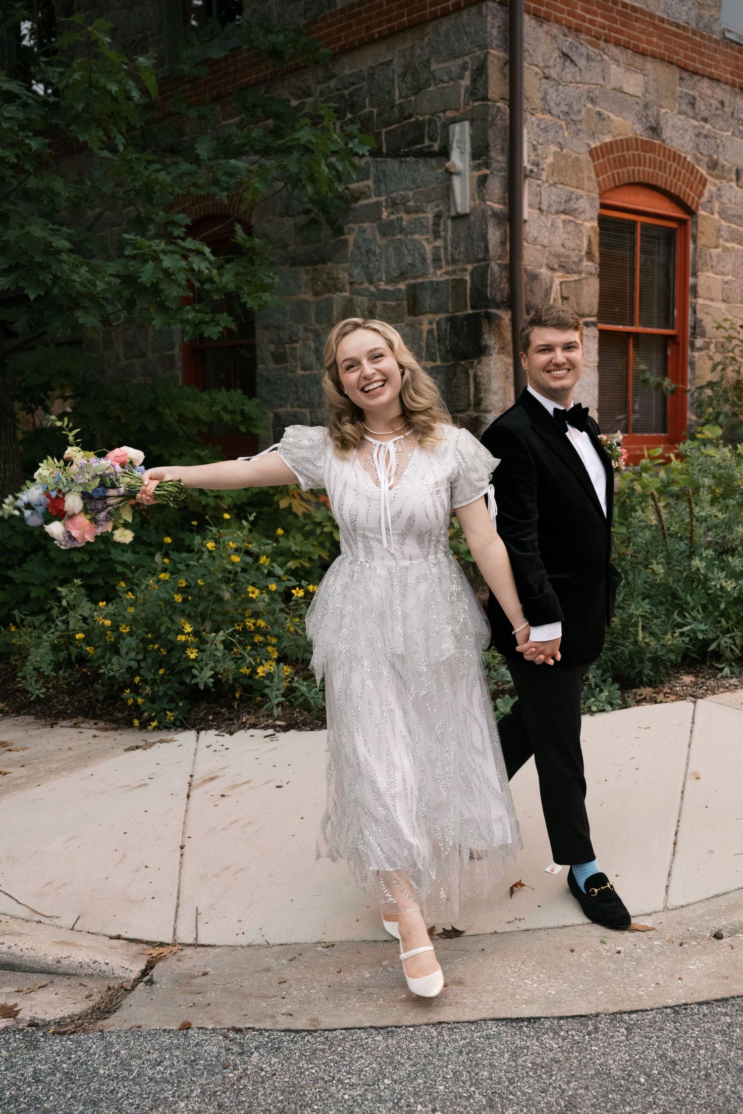 A couple poses joyfully outside a historic stone building with flowers.