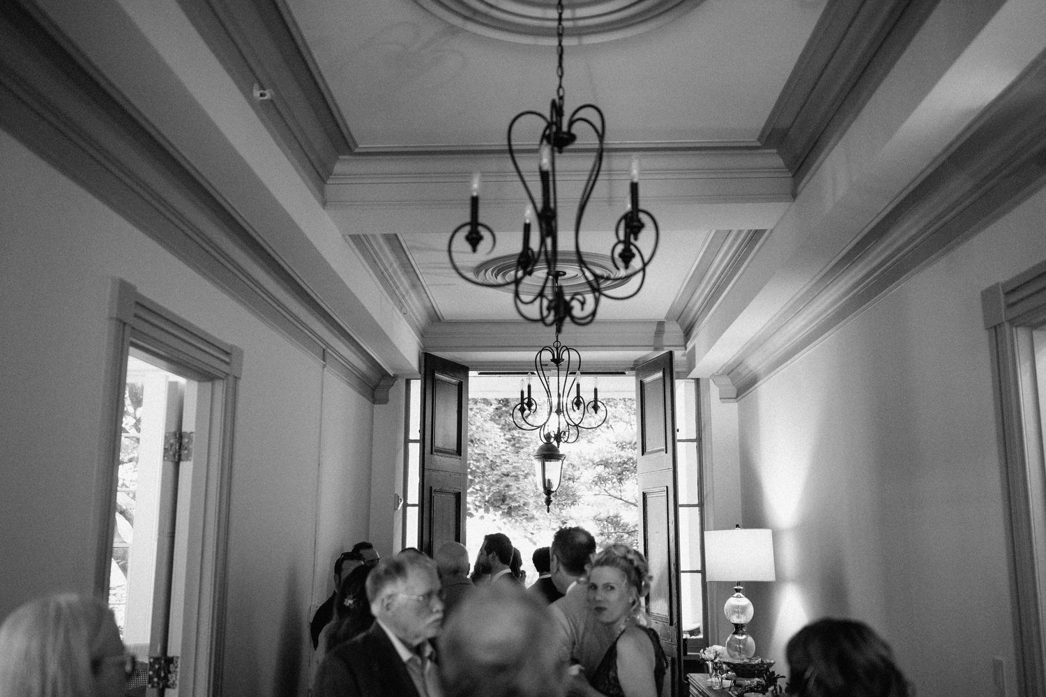 Elegant chandelier hangs in a bright white hallway during a gathering.