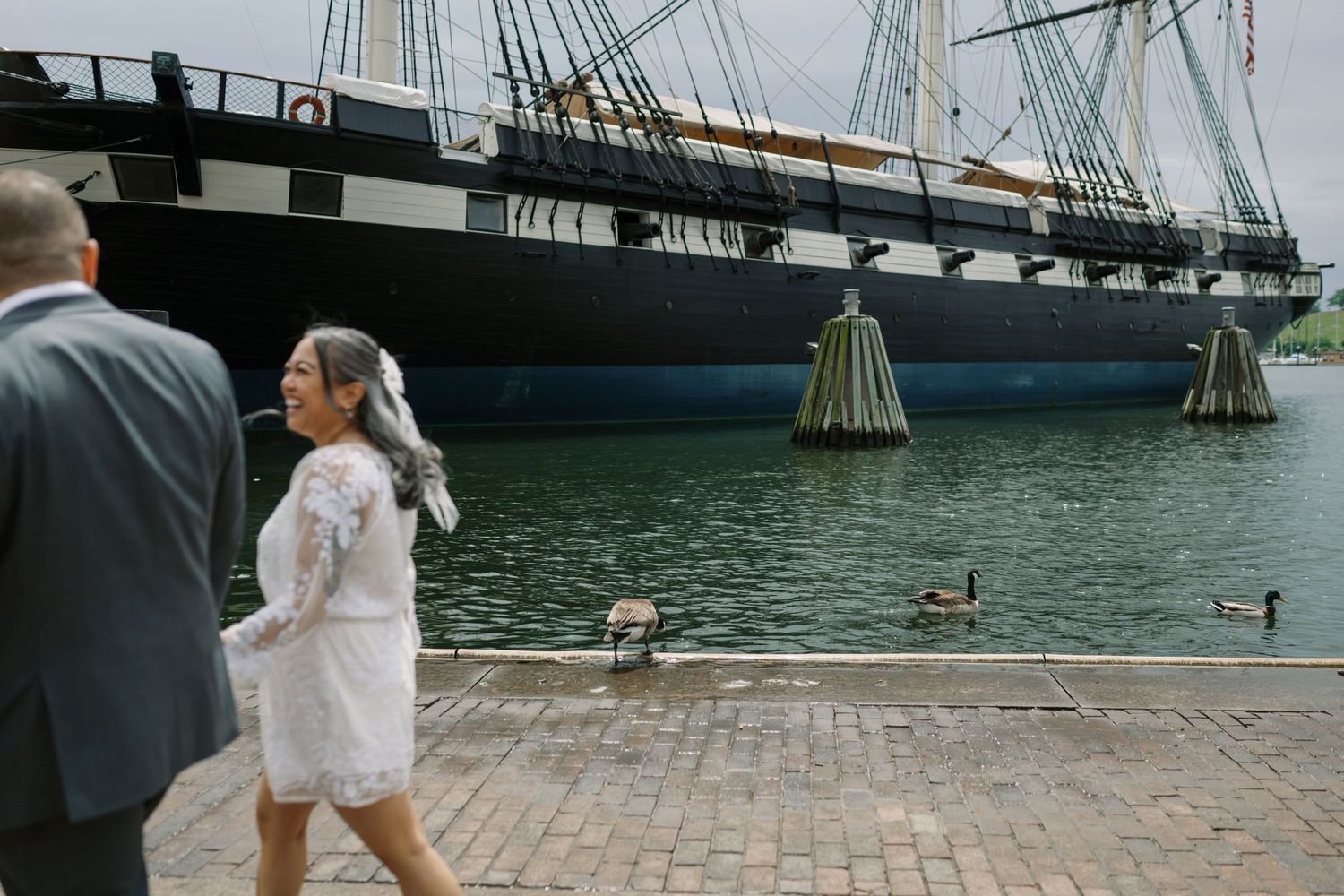 Two people stroll along a waterfront boardwalk near a historic tall ship docked at a maritime museum.