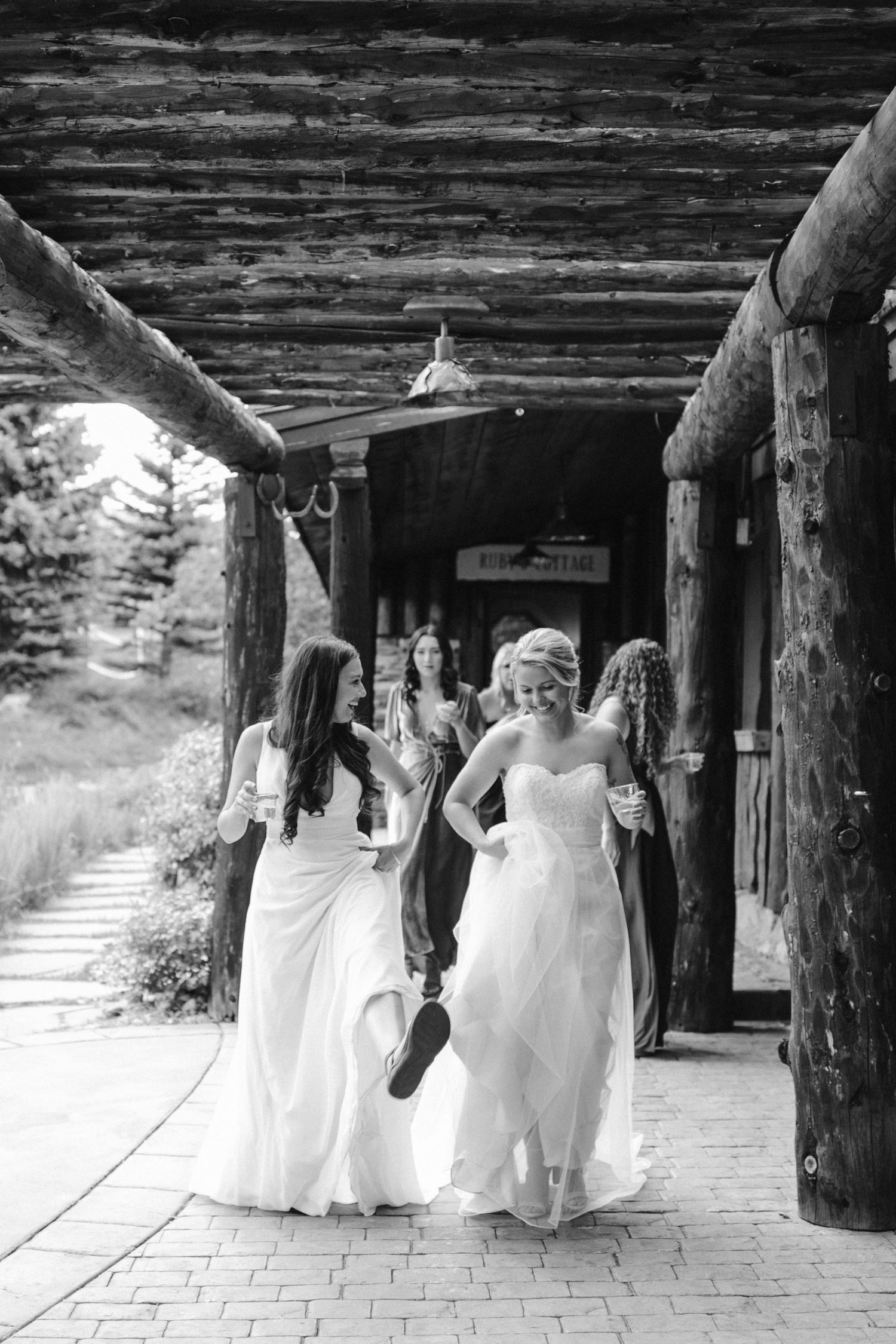 Two people in wedding dresses walk together under a rustic log cabin porch.