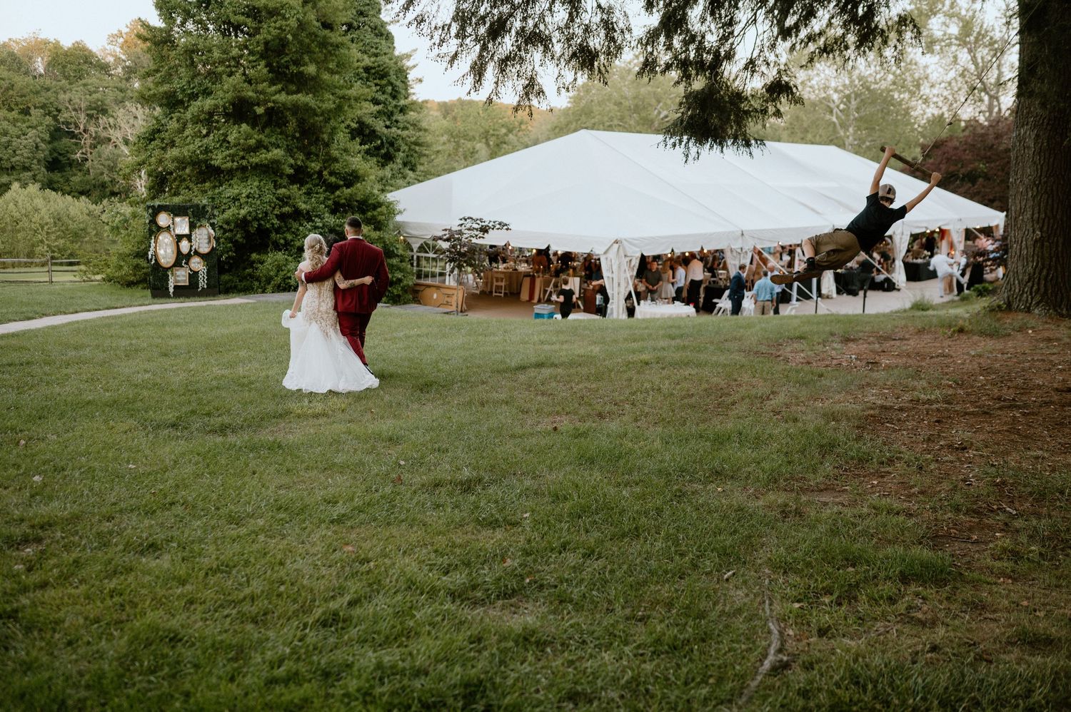 An outdoor wedding celebration with white tents set up on a grassy lawn near trees.