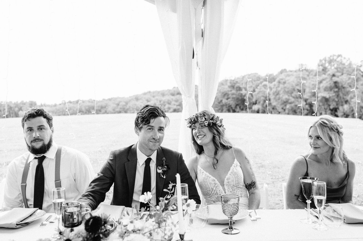 Candid reception table moment under white tent with guests enjoying drinks and conversation at an outdoor wedding.