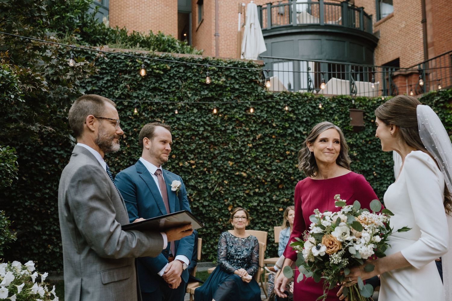 An intimate outdoor wedding ceremony takes place in a garden setting with ivy-covered walls and floral arrangements.
