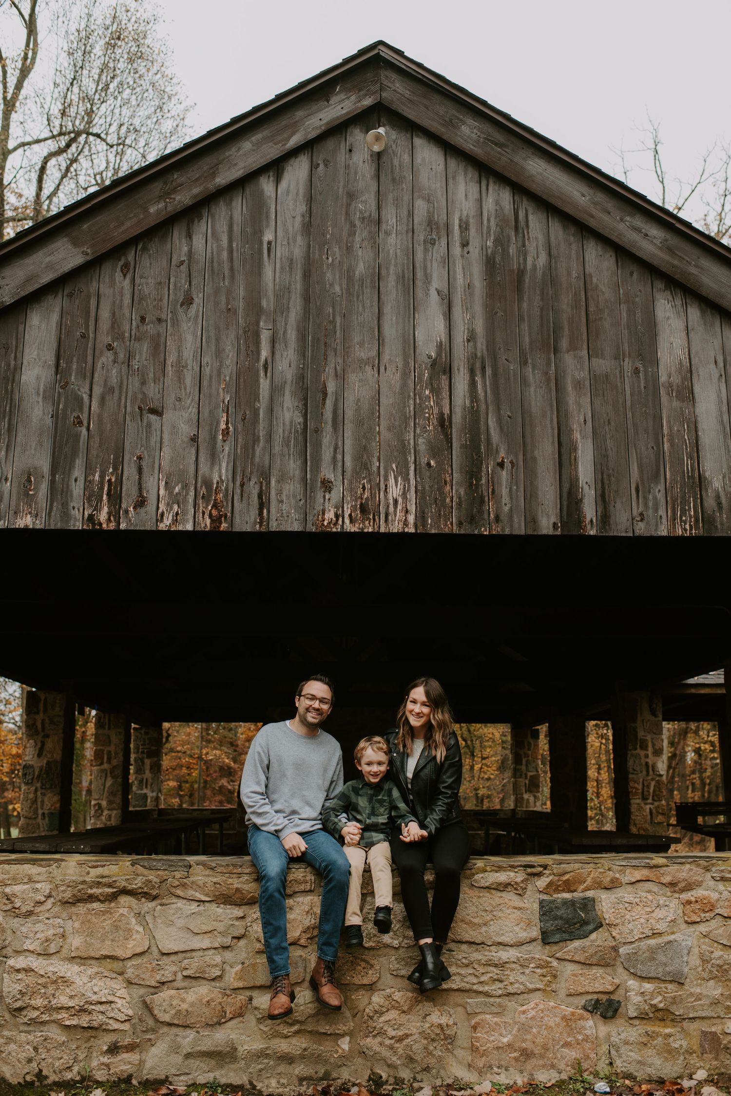Family sitting together under a rustic wooden shelter with stone base during fall season.
