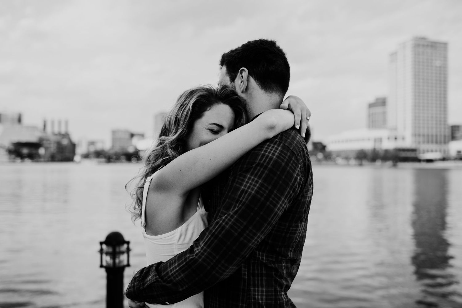 Romantic couple embracing by the waterfront with city skyline in black and white.