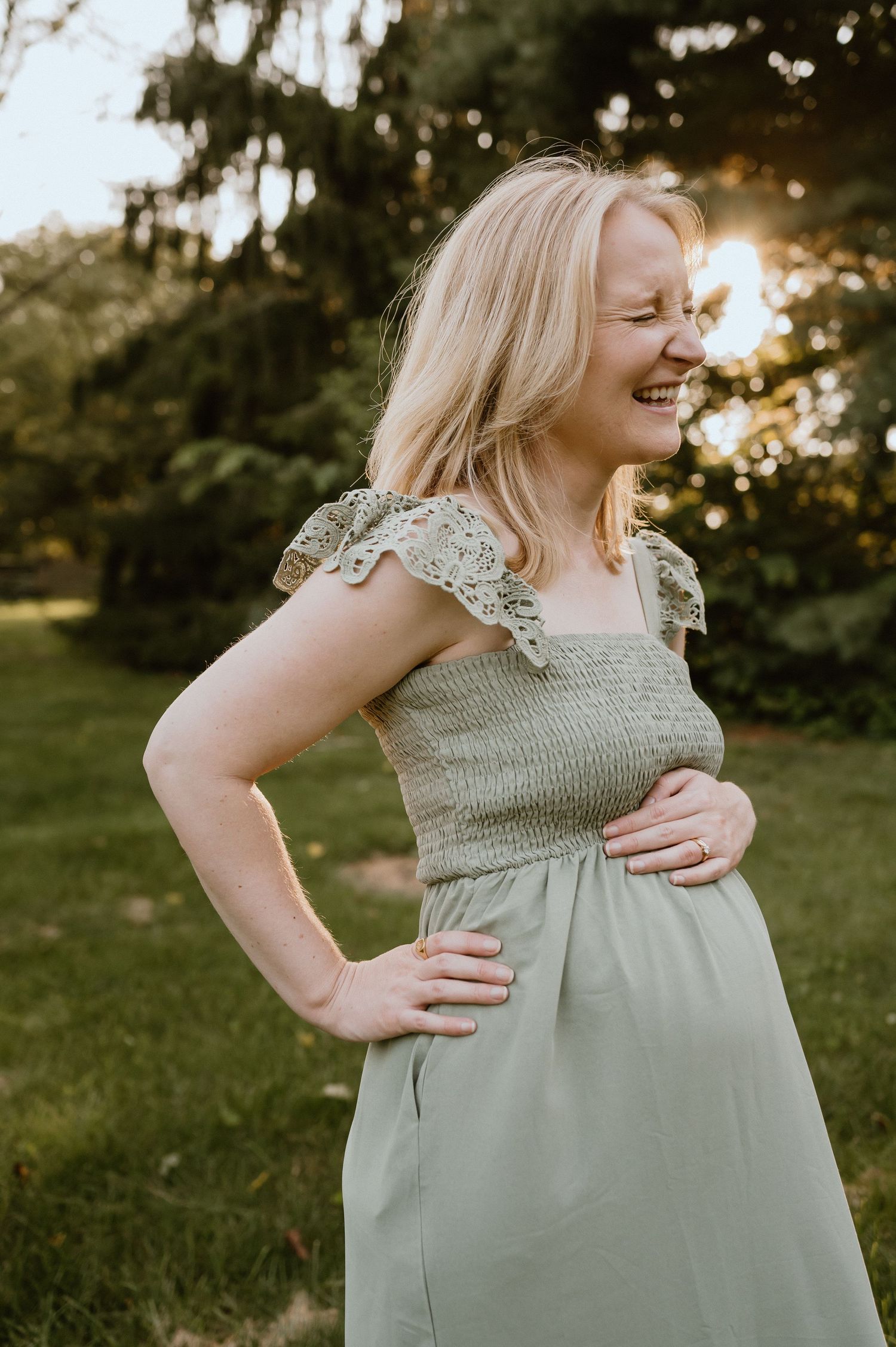 A woman in a sage green dress with lace details poses outdoors at sunset with a joyful expression.