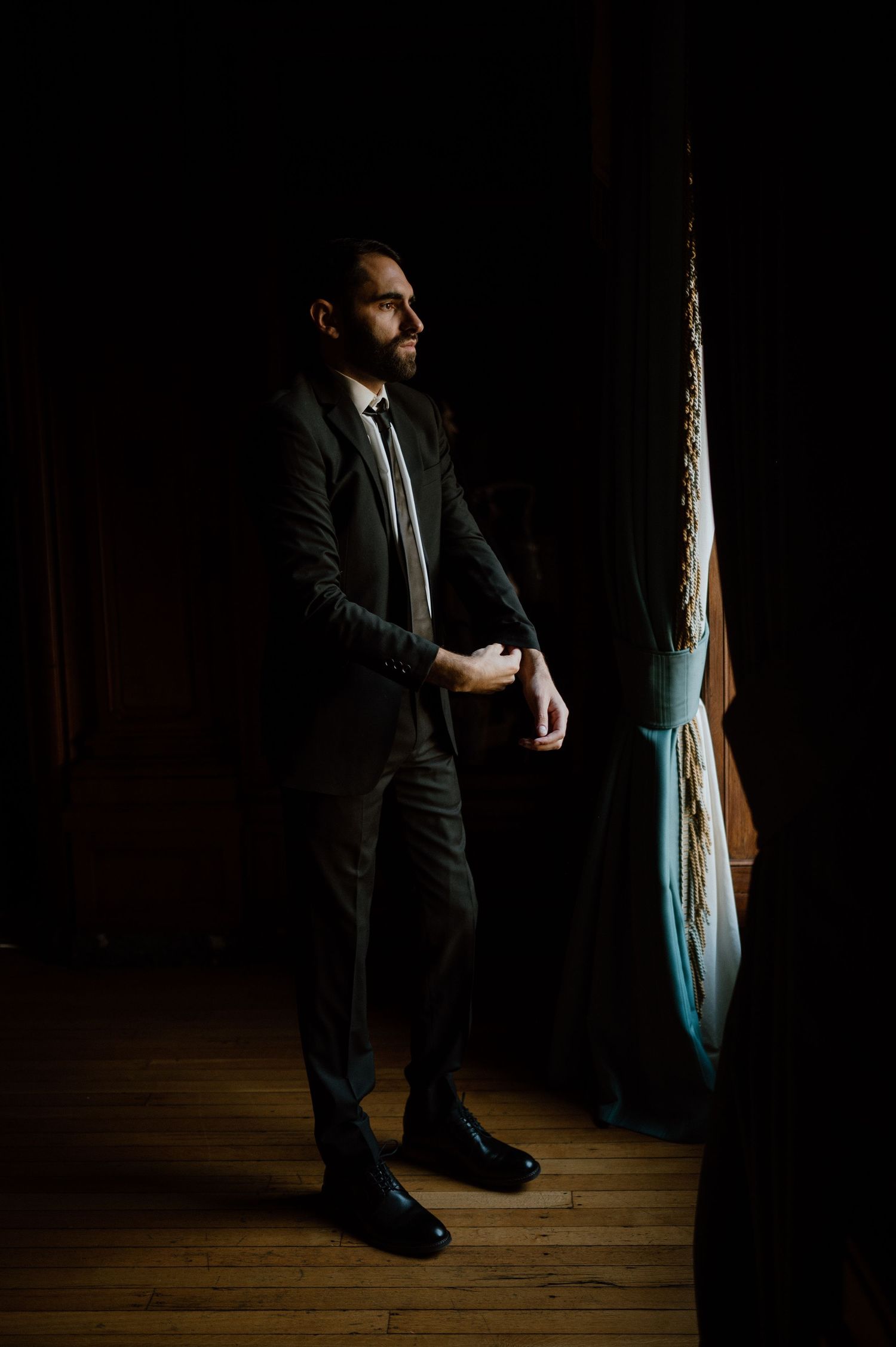 Dramatic portrait of someone in a dark suit standing by curtains in moody lighting.