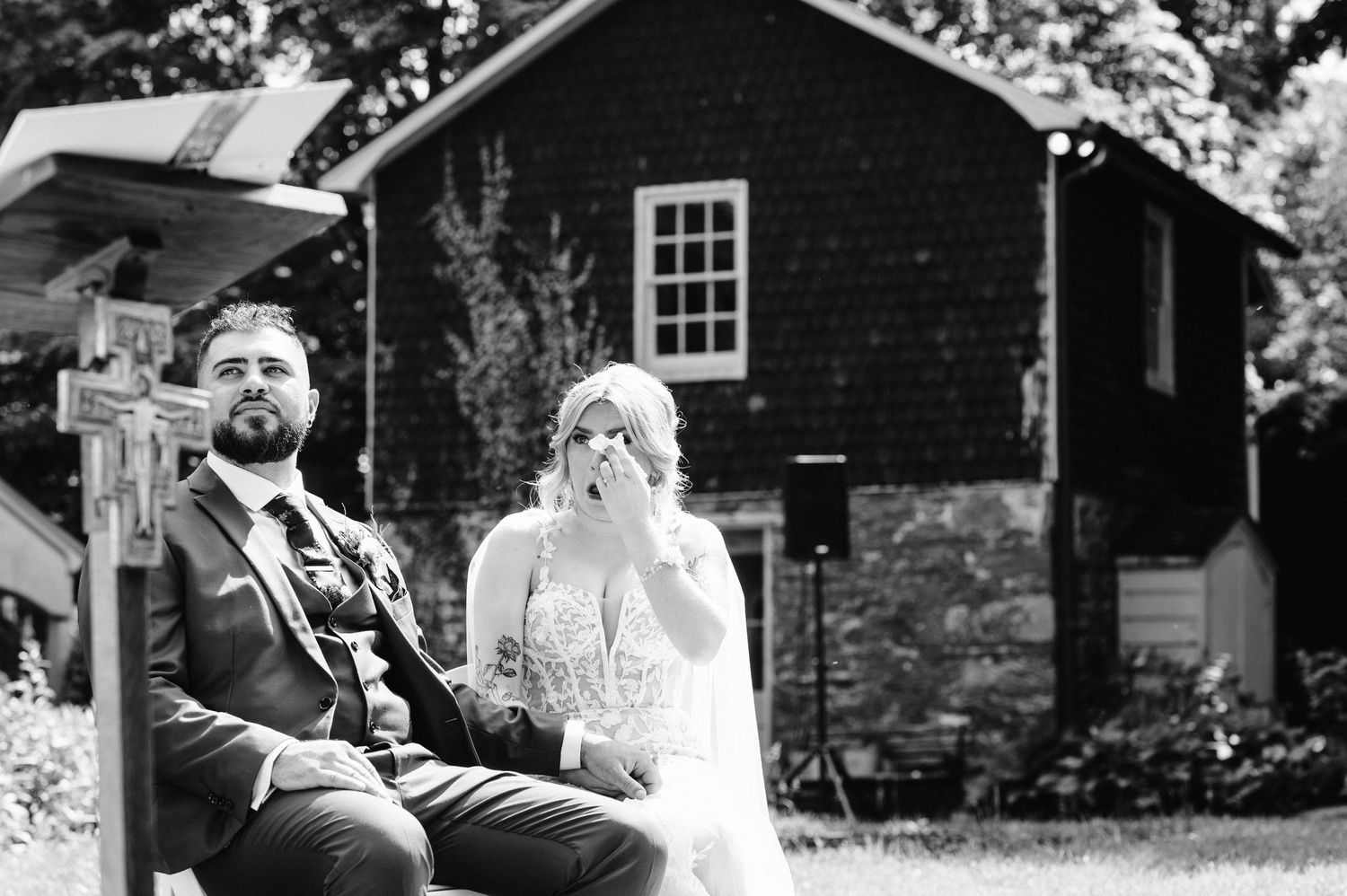 A black and white wedding portrait of a couple seated outdoors near a rustic building.