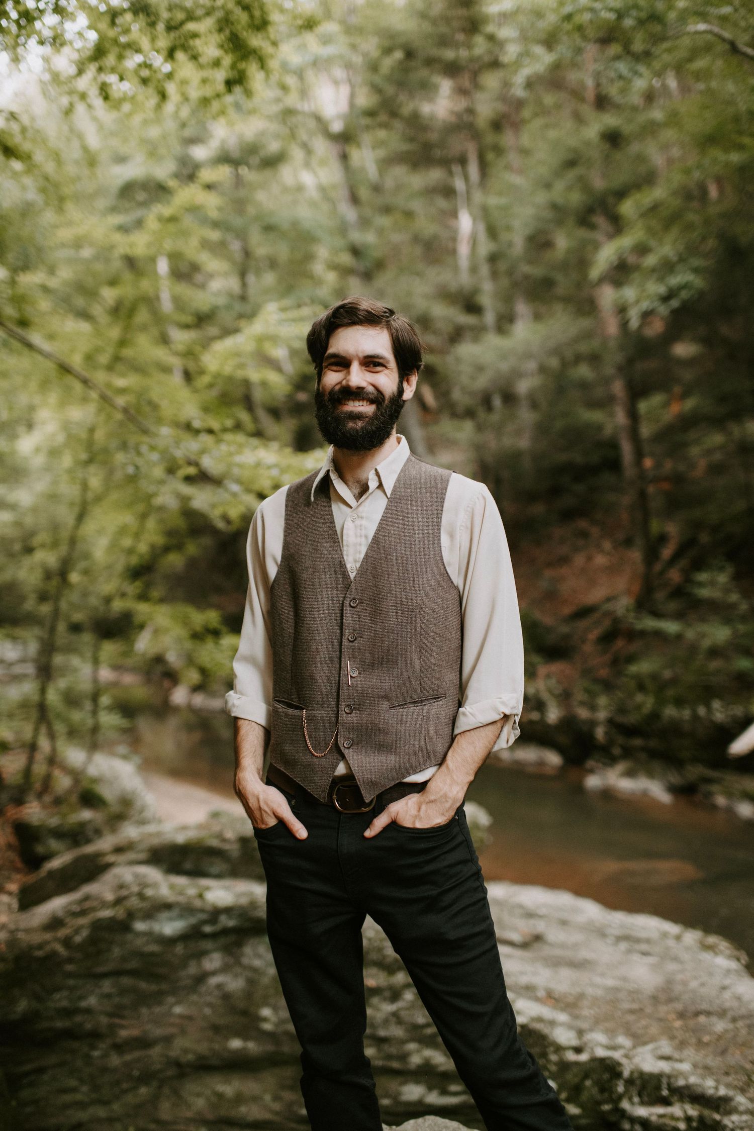 A groom in casual wedding attire stands on rocks by a forest stream with a natural smile.