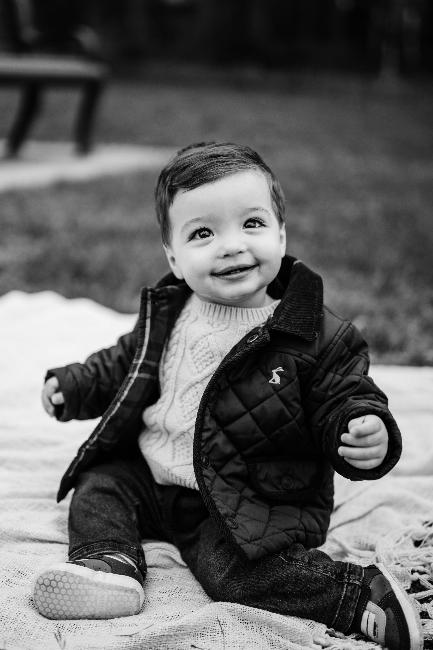 Black and white portrait of baby in quilted jacket sitting on blanket outdoors.